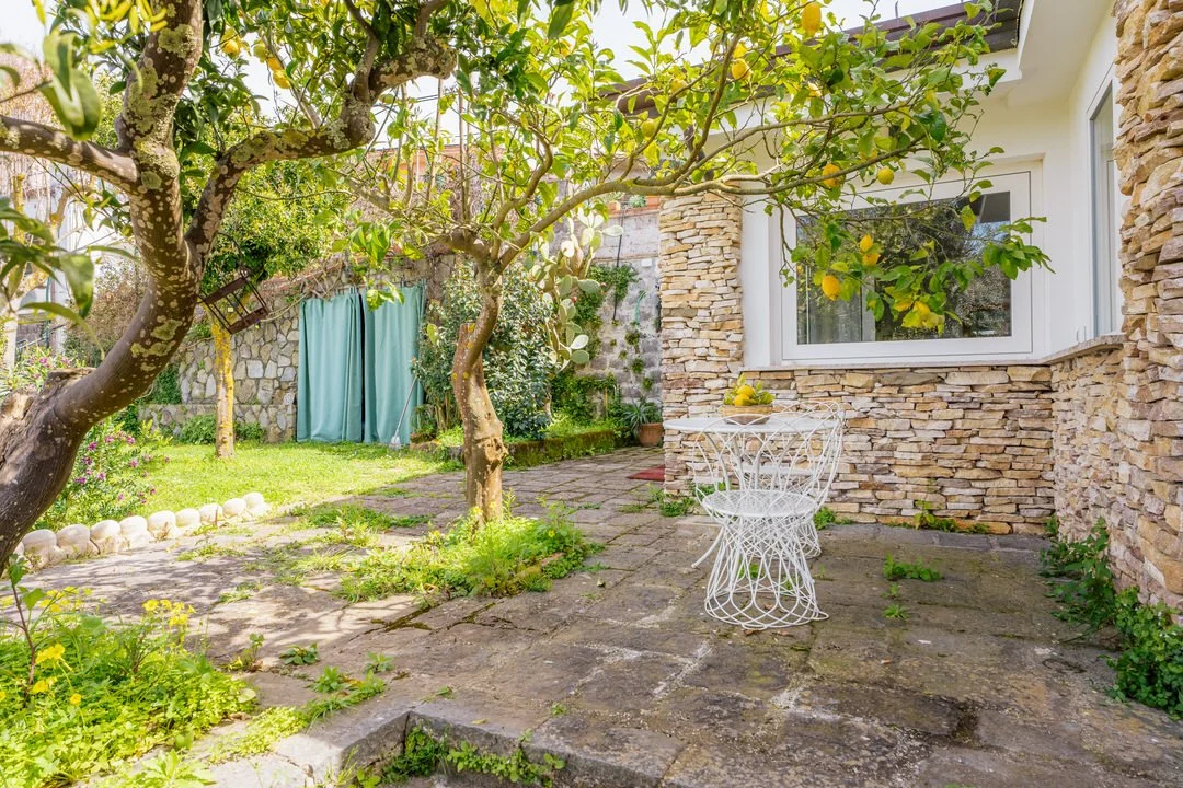 A cozy backyard patio with stone flooring, surrounded by greenery and trees, featuring a white wire table and chairs, a lemon tree with ripe lemons, and a house with a stone wall and large window.