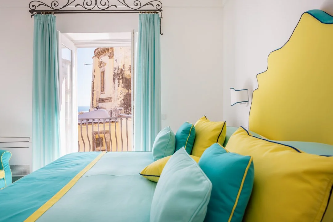 Hotel room with turquoise and yellow bedding, open door leading to balcony with view of an old building and the ocean