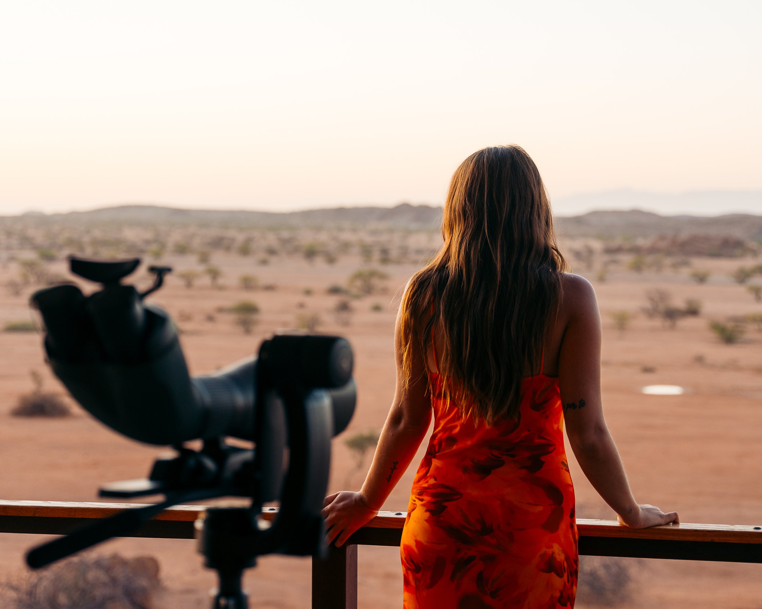 A woman with long brown hair and tattoos on her arms, wearing a red and orange tie-dye dress, gazes at a desert landscape with scattered bushes at sunset. A camera is mounted on a tripod in the foreground.