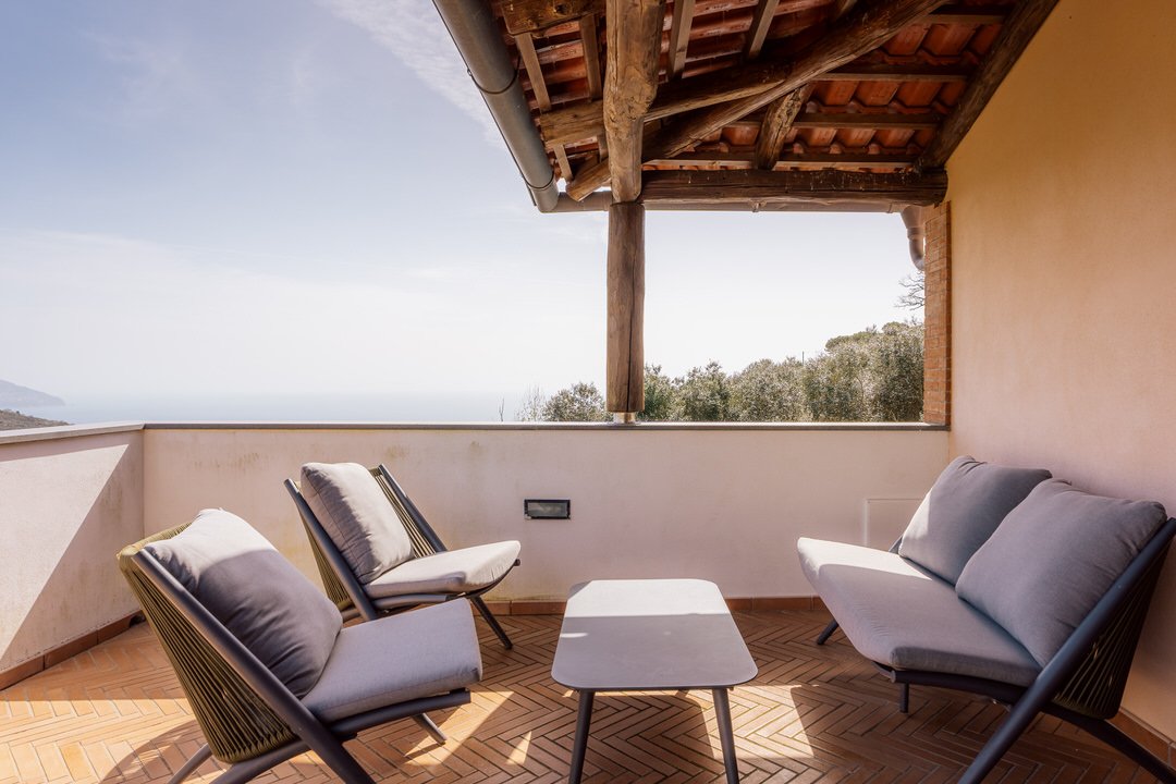 Outdoor balcony with two armchairs and one sofa, overlooking a landscape with trees and distant hills, covered by a wooden roof.