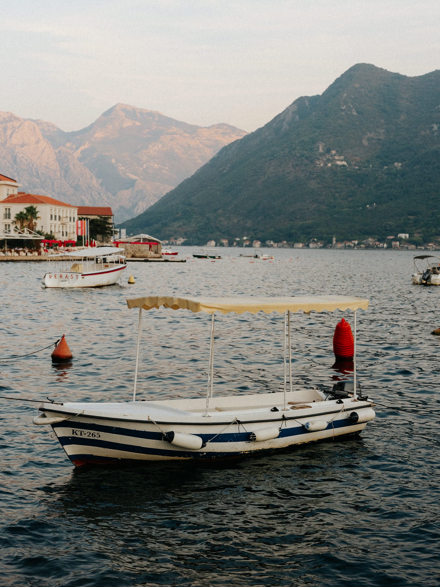 A small white boat with a black stripe and registration number 'KT-265' floating on a dark body of water with boats and mountains in the background.