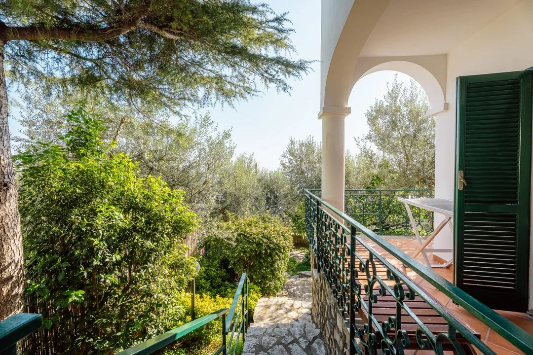View from a balcony overlooking a lush garden with trees and shrubs, with an open door and a white folding chair on the balcony