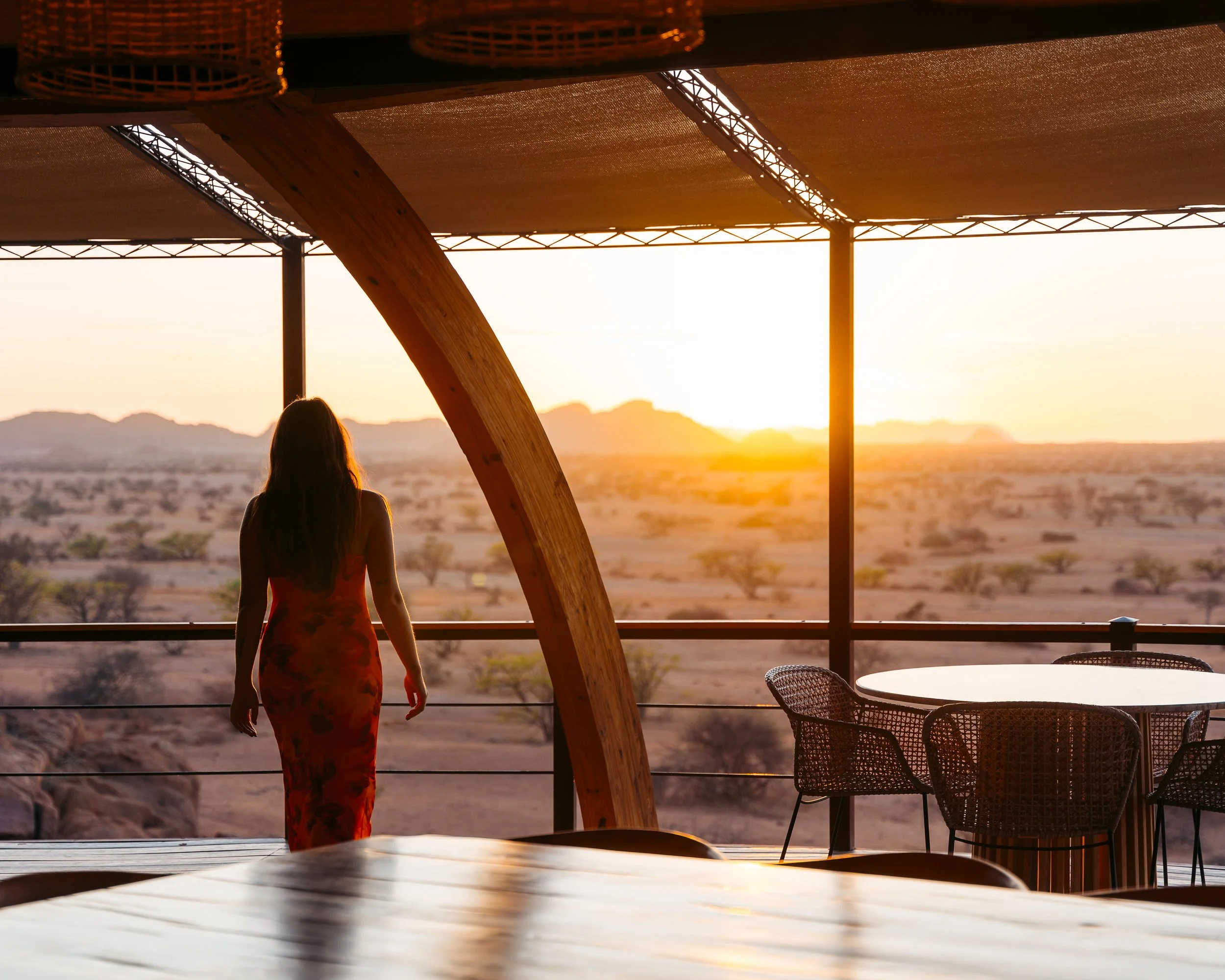 A woman standing on a balcony overlooking a desert landscape during sunset, with a dining table and chairs nearby.