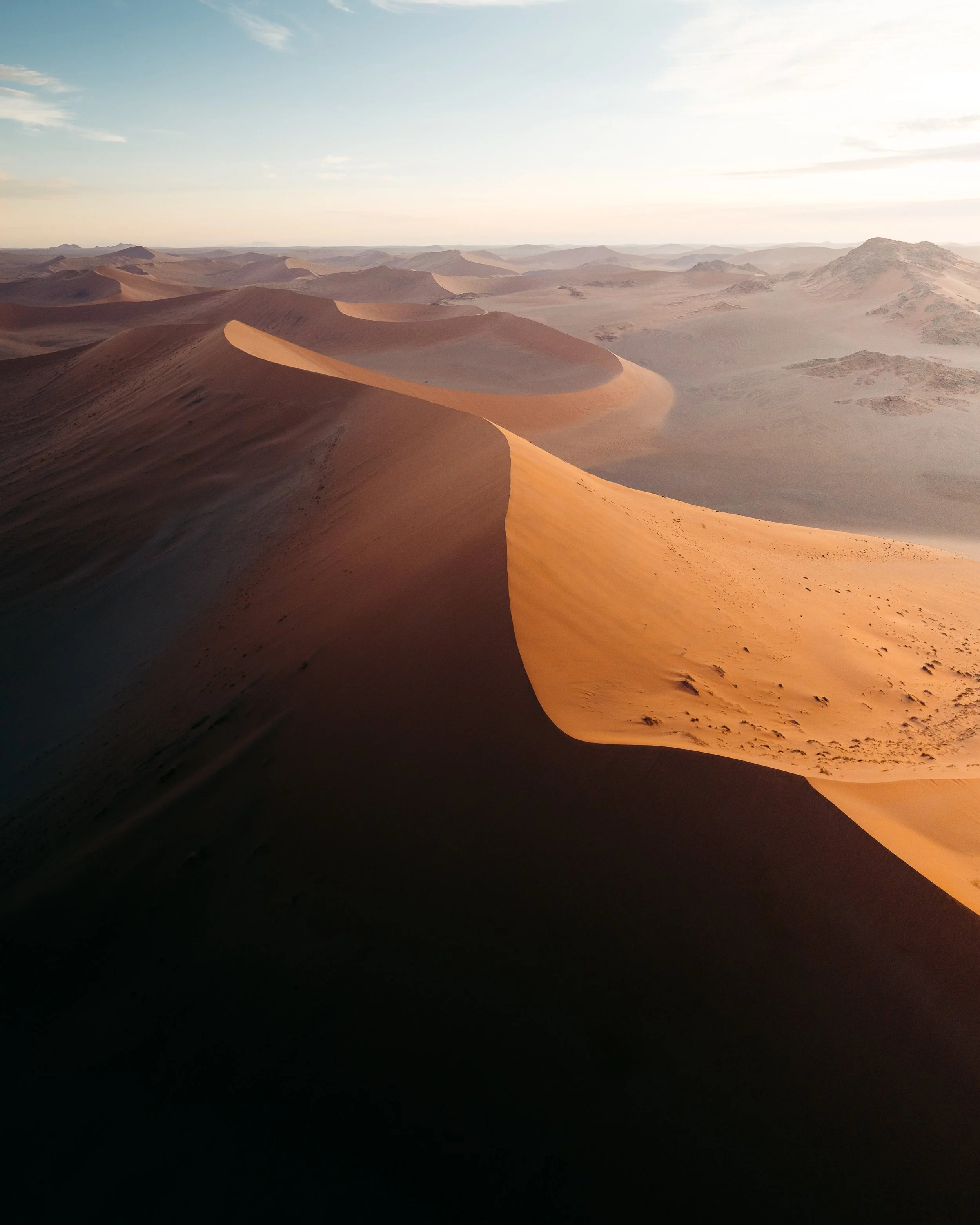 Aerial view of vast sand dunes in a desert at sunset with a sky filled with clouds.