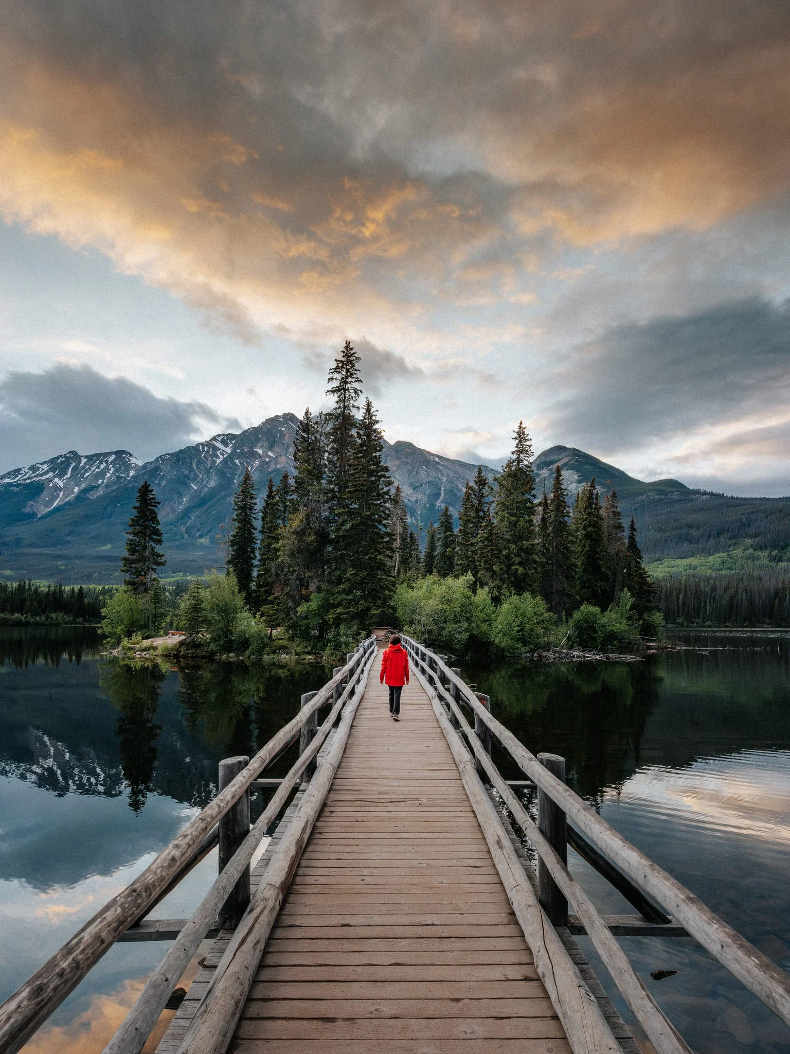 A person in a red jacket walking on a wooden bridge over a calm lake, surrounded by tall pine trees with mountain peaks in the background and a cloudy sky above.