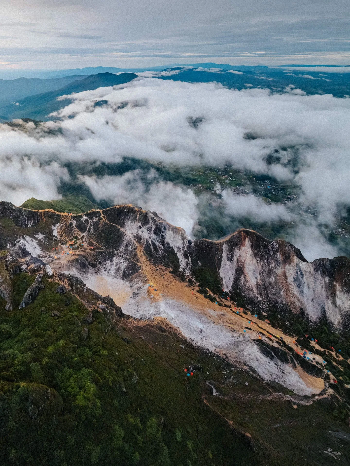 Mountain landscape with tents near rocky peaks, clouds covering the valley below, and distant mountains under a cloudy sky.