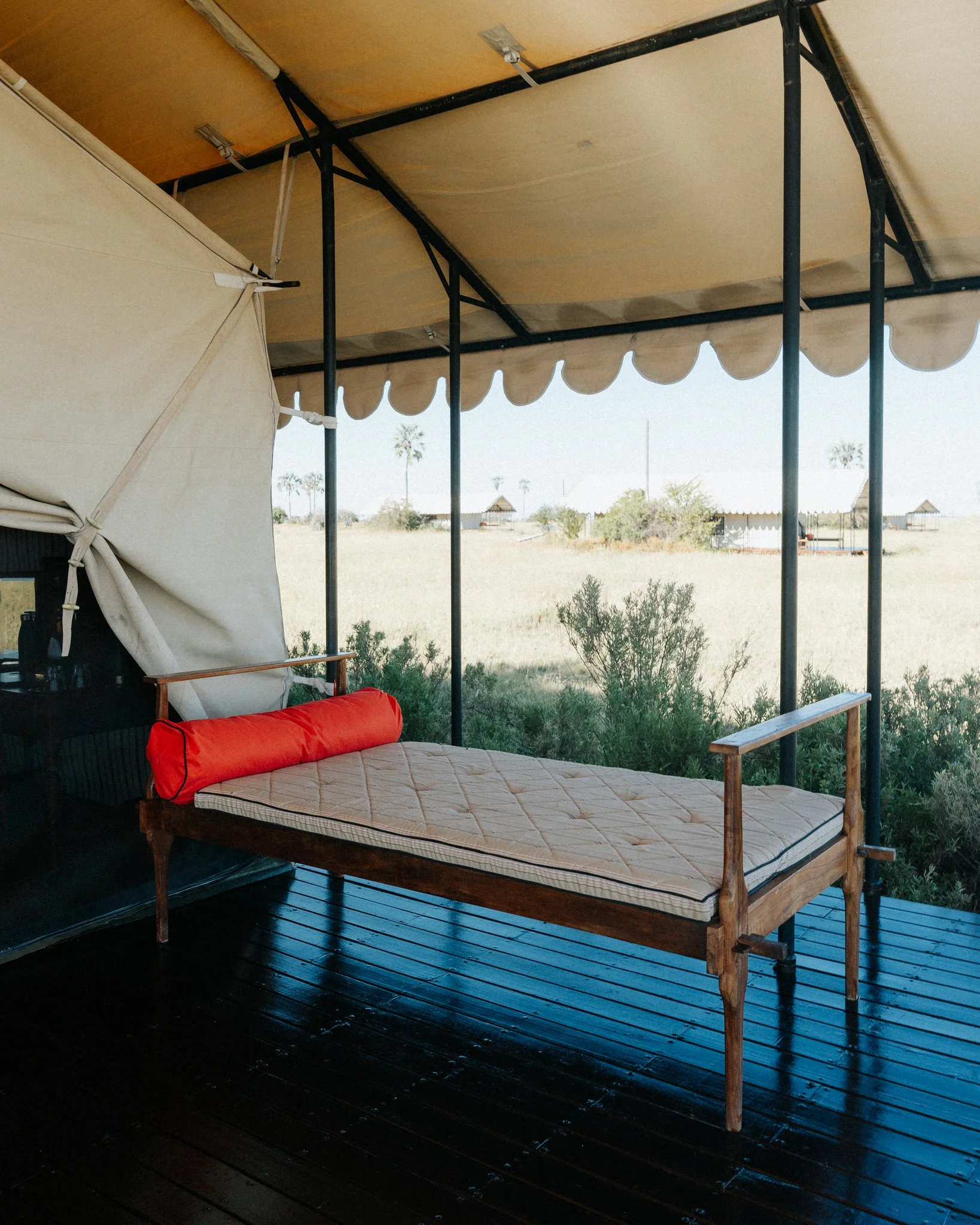 Outdoor patio with a black wooden floor, a wooden daybed with a mattress and a red bolster pillow, under a beige canopy with scalloped trim, and a view of a dry landscape with distant palm trees and small structures.