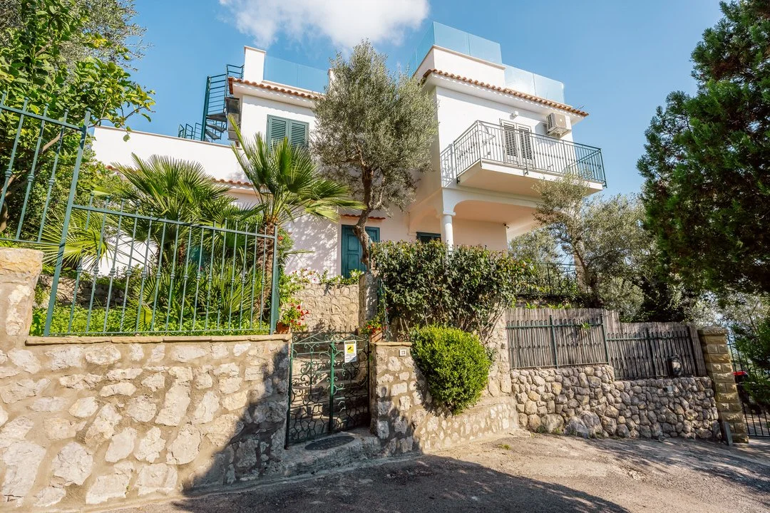 A white multi-story house with balconies, surrounded by trees and greenery, with a stone wall and metal fence in the foreground.