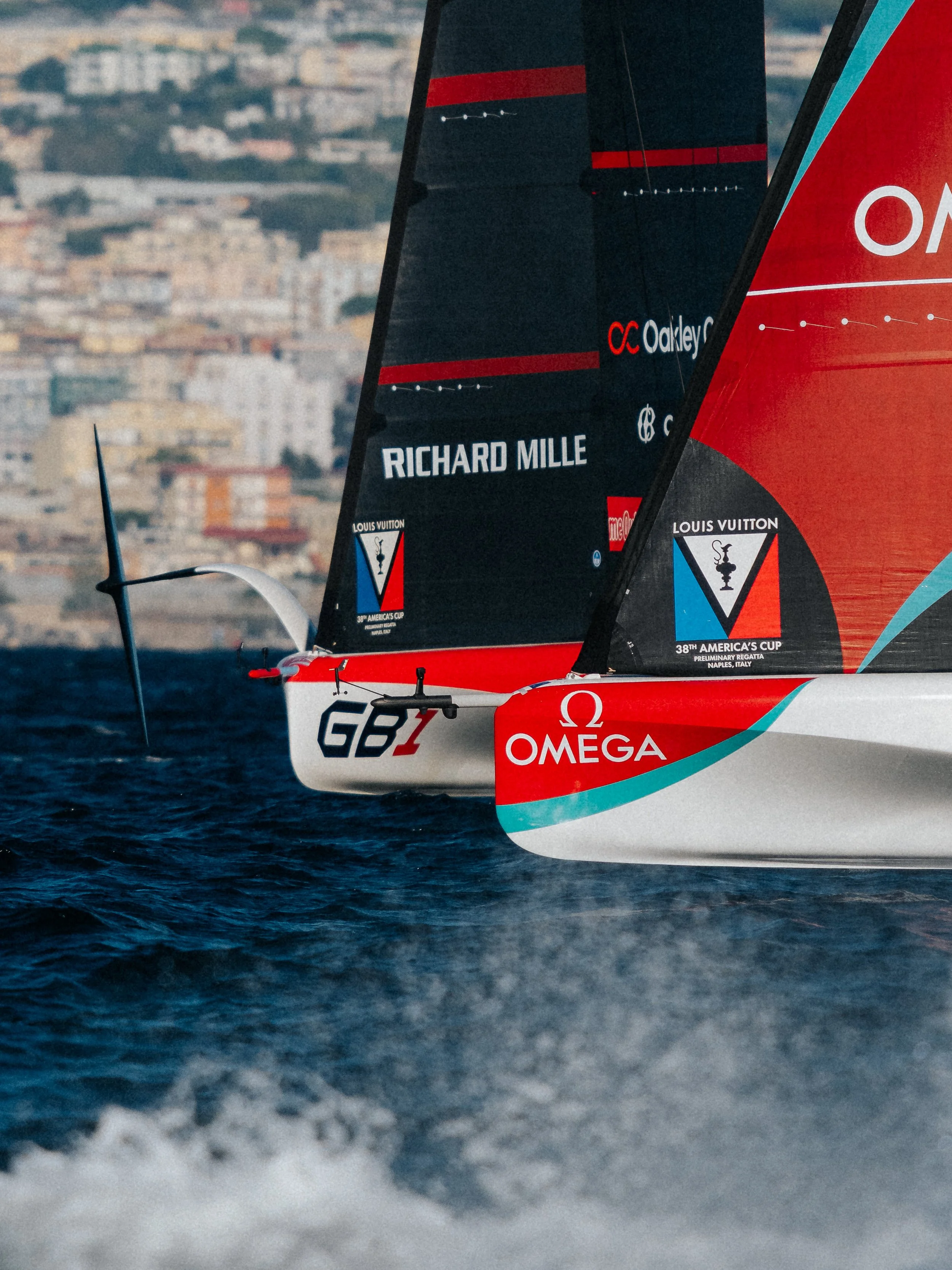 Close-up of a racing sailboat's sail with sponsor logos, including Richard Mille and Louis Vuitton, moving through the water near a city shoreline.