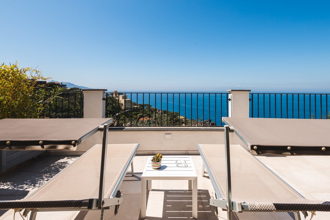 A balcony with two reclining lounge chairs, a small white table with a plant and glass, overlooking a coastal view of the ocean and distant hills under a clear blue sky.
