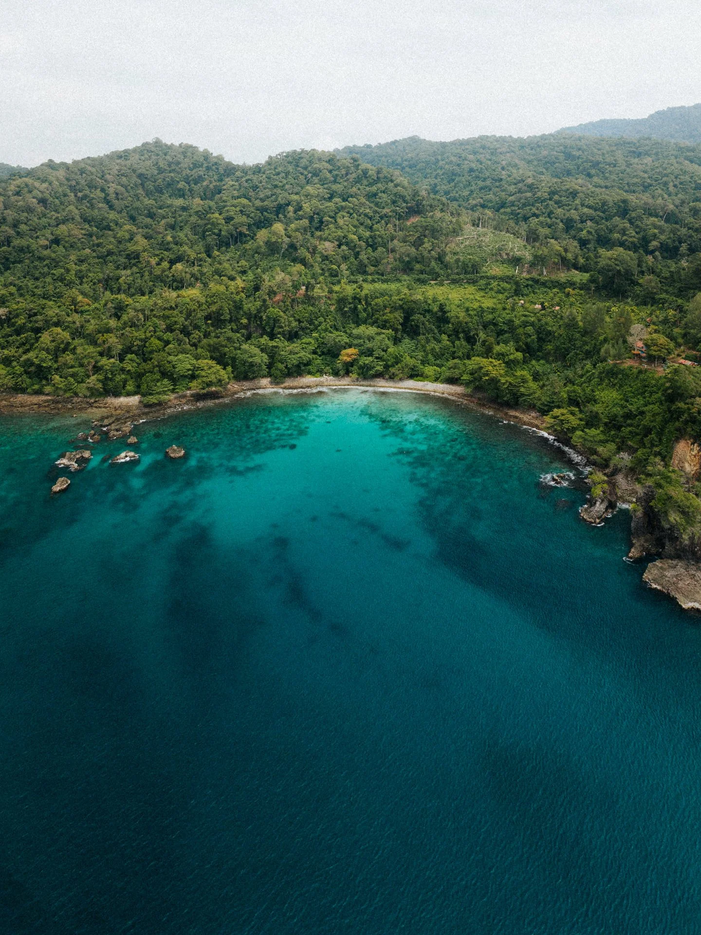 Aerial view of a tropical bay with clear blue water, surrounded by lush green mountains and rocky shoreline.