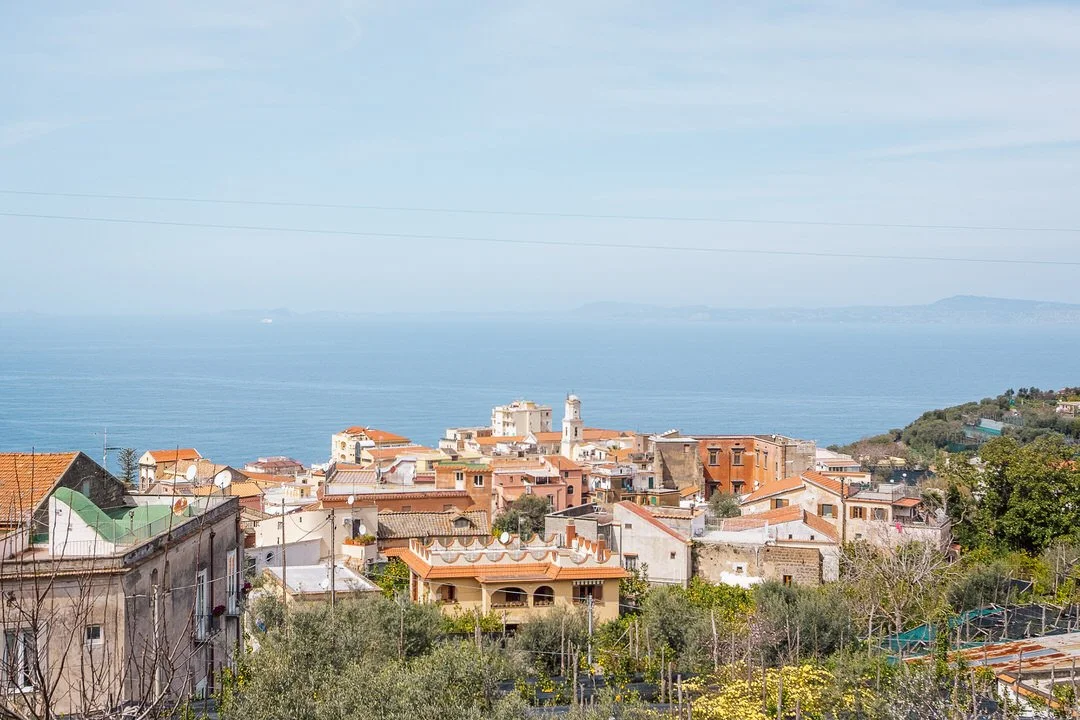 View of a coastal town with houses and buildings near the shoreline, overlooking a large body of water under a partly cloudy sky.