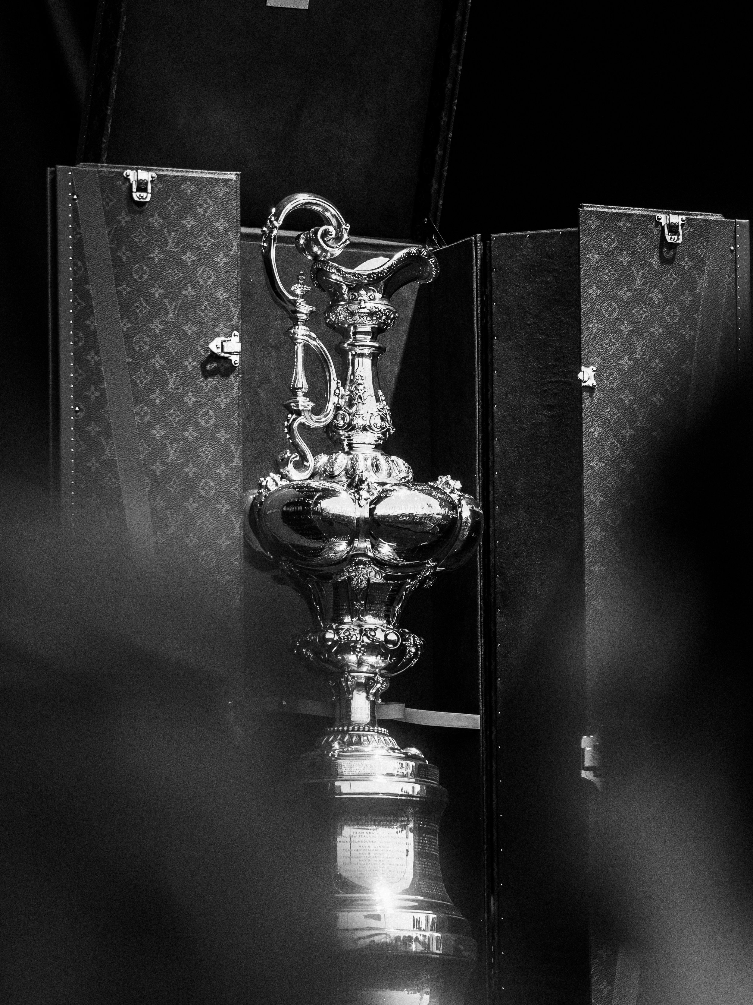A silver trophy or award with ornate design, placed on a reflective surface with its reflection visible.