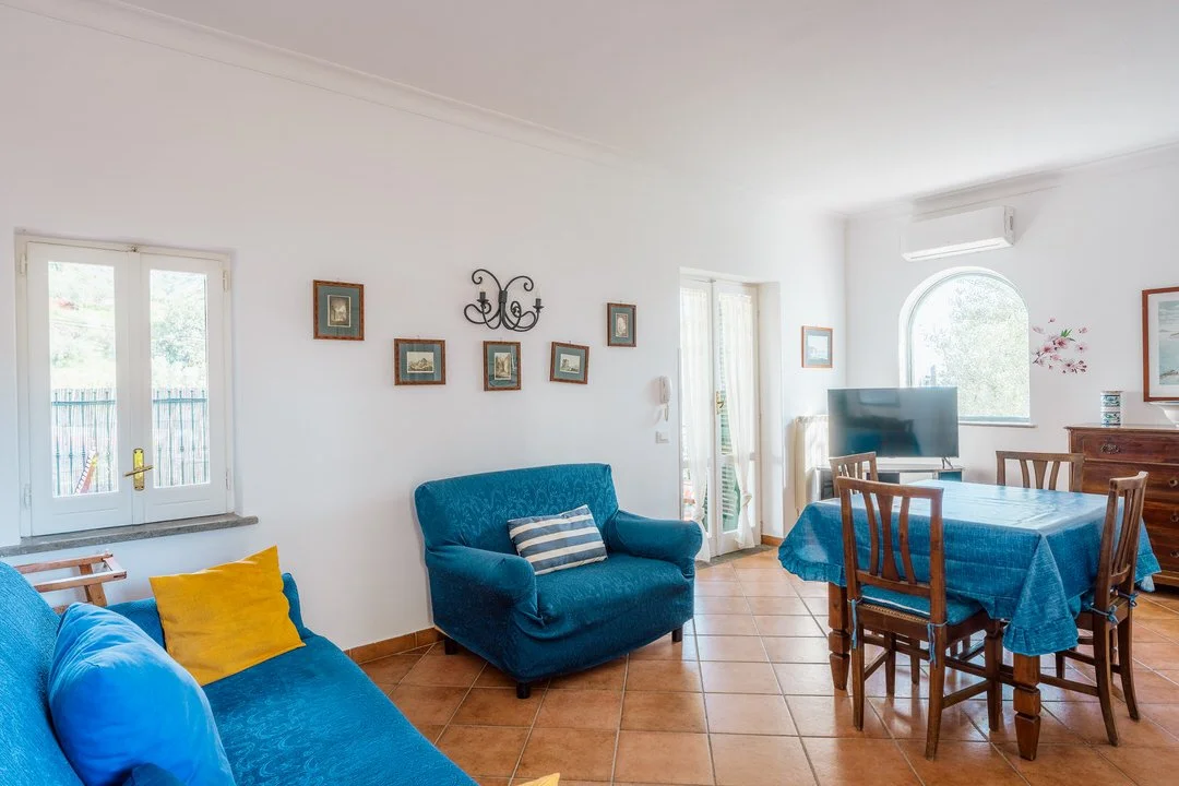 Living room with blue sofas, wooden dining table with chairs, a television, windows, framed pictures, and terracotta tile flooring.