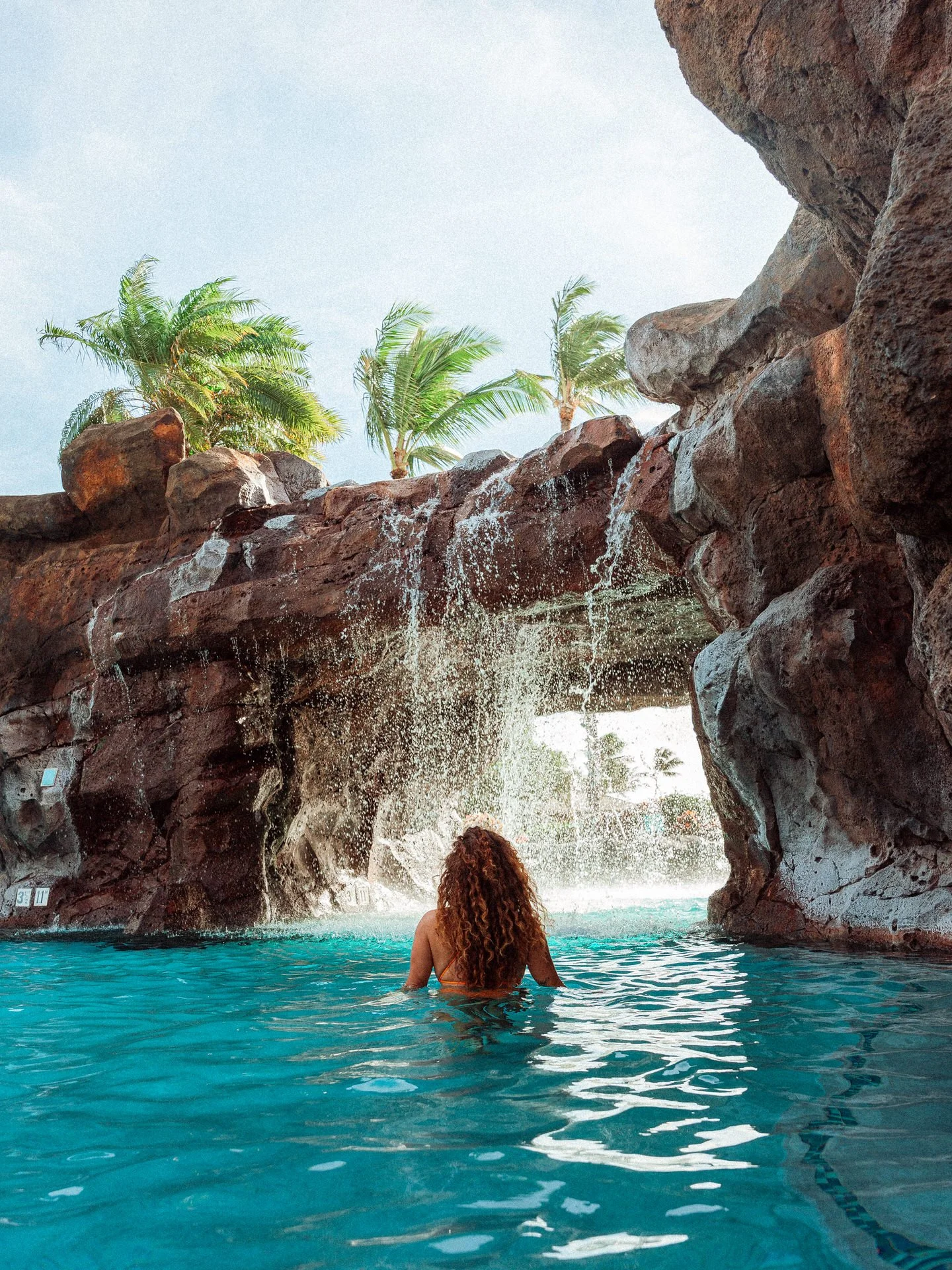 A woman with curly hair swimming in a pool with a waterfall feature, surrounded by rocky structures and palm trees in the background.