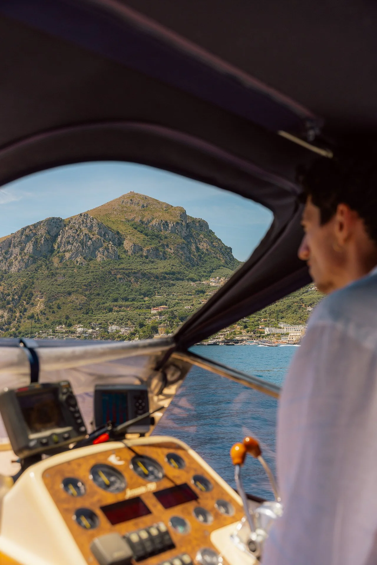 View from inside a boat's cabin showing a man looking at a mountain across the water, with the boat's control panel in the foreground.