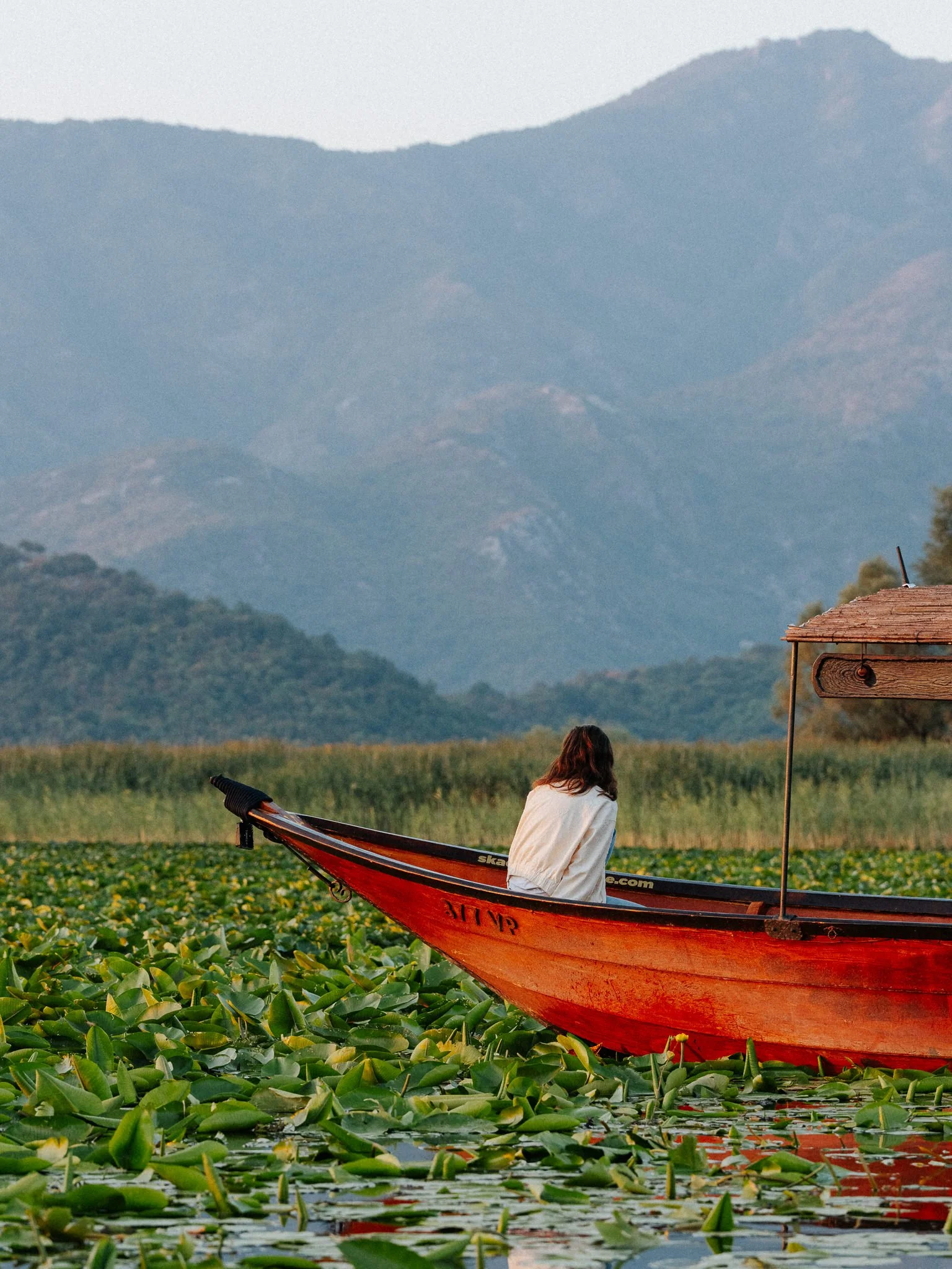 A person with long hair sitting on a red boat in a waterbody covered with green aquatic plants, with mountains in the background and a cloudy sky above.
