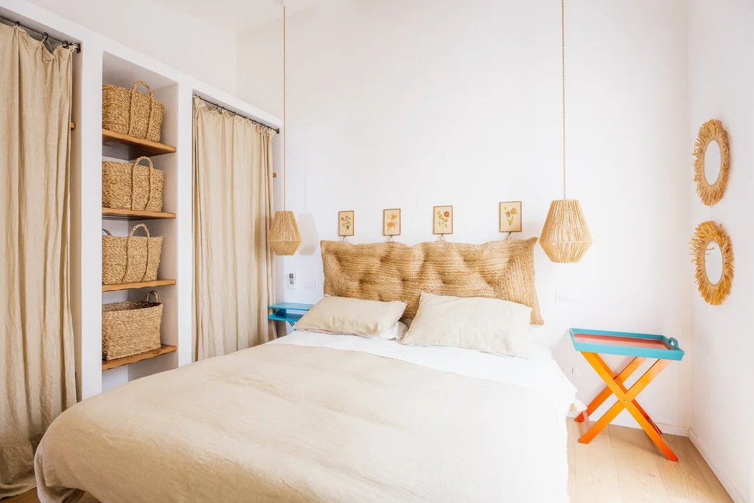 A bedroom with a white bed, beige pillows, woven headboard, two hanging rattan pendant lights, framed botanical art, a blue nightstand, and a colorful folding table, with woven baskets on open shelves and circular wicker wall decor.