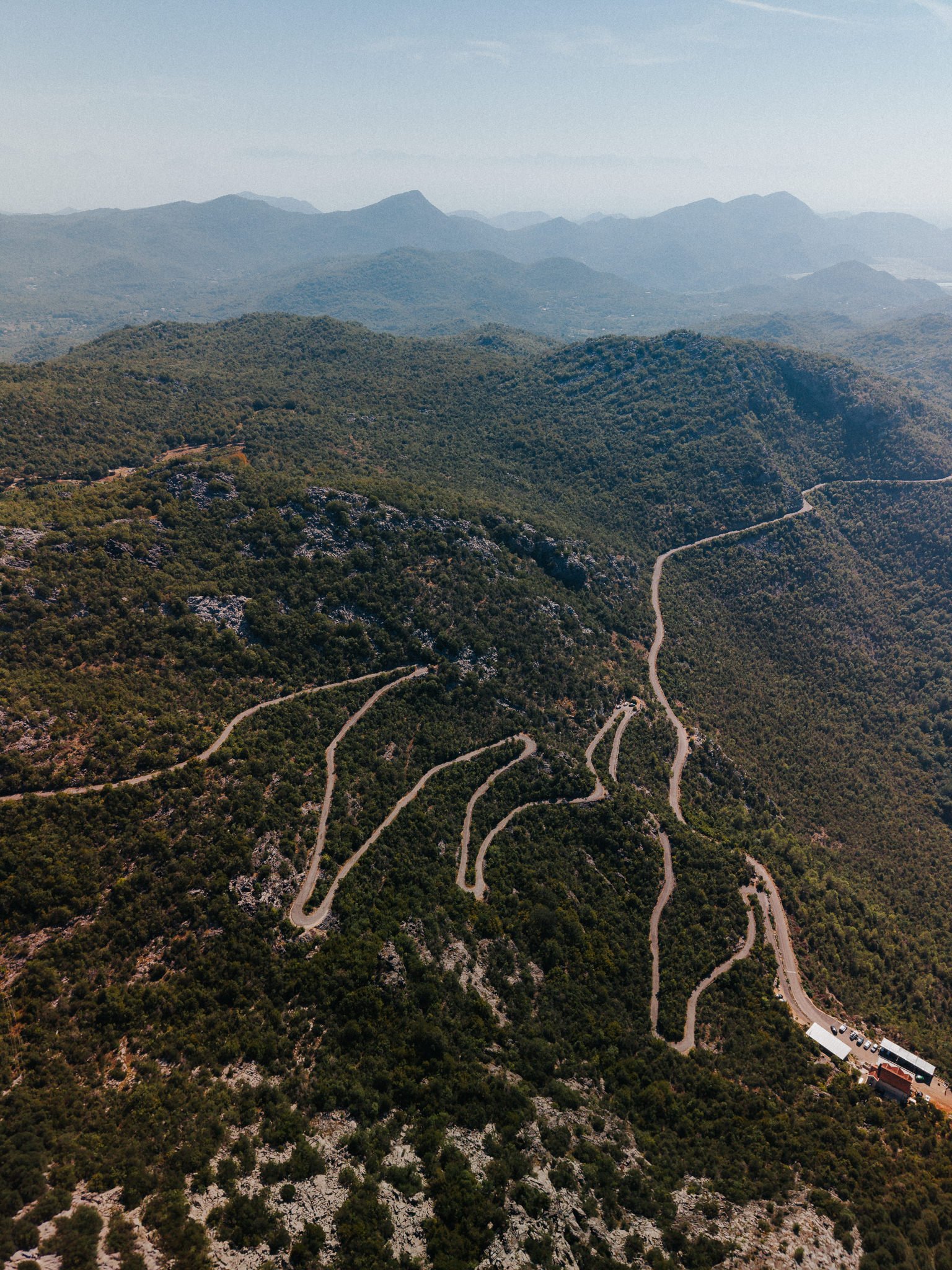 A winding mountain road runs through dense green forest, with mountain peaks visible in the distance, under a clear sky.