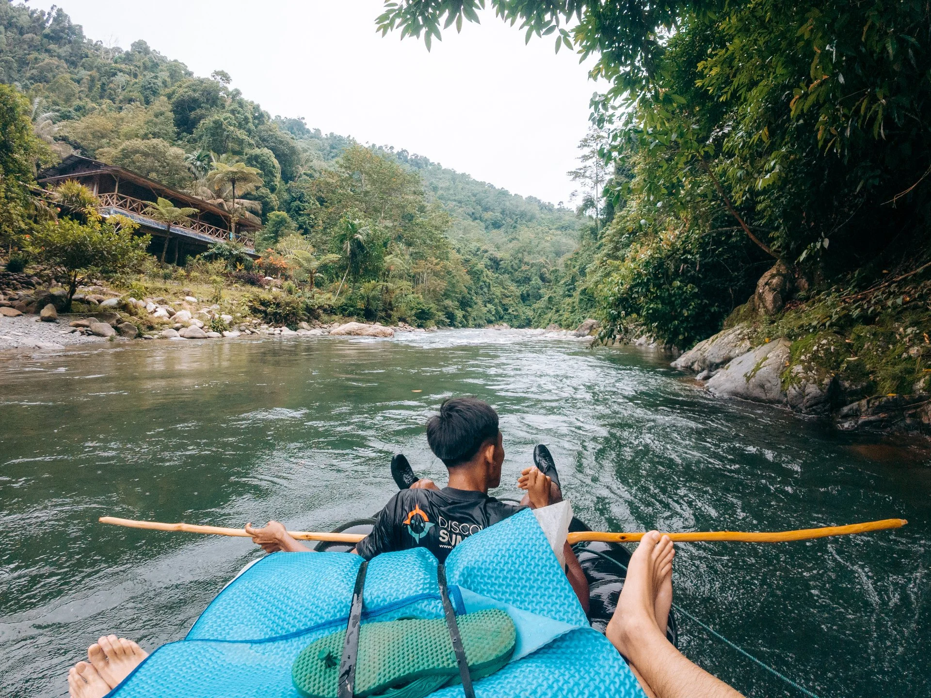 People rafting down a river in lush green forest with rocky banks and a wooden building on the hillside.