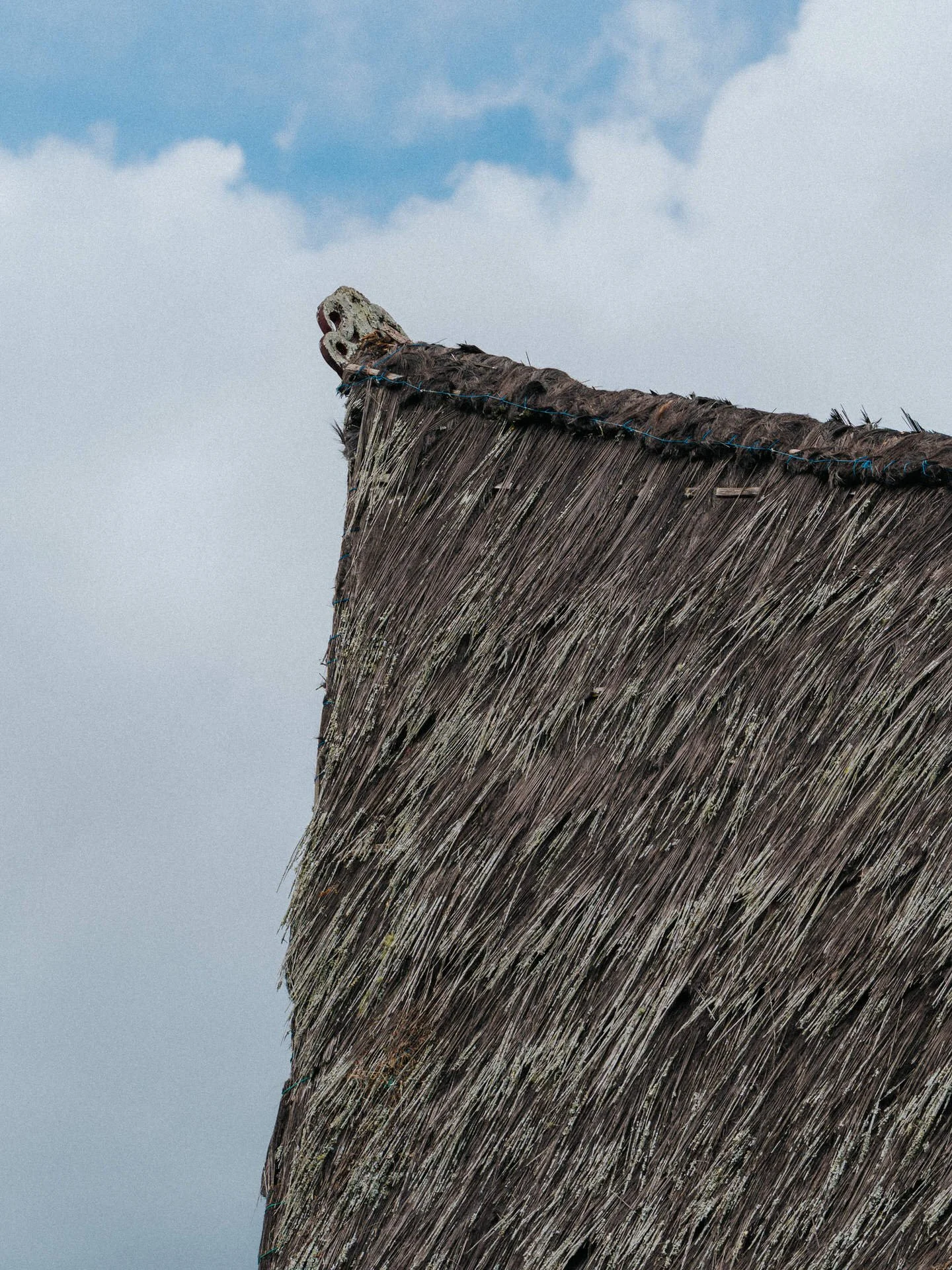 Close-up of a thatched roof with a carved wooden figure at the point, against a background of sky with clouds.