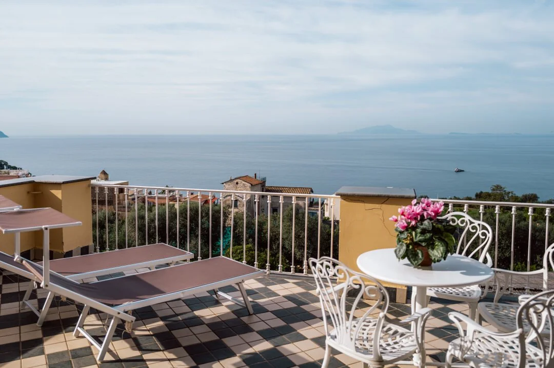 Balcony with lounging chairs and a table with pink flowers overlooking a seaside view with water and distant islands