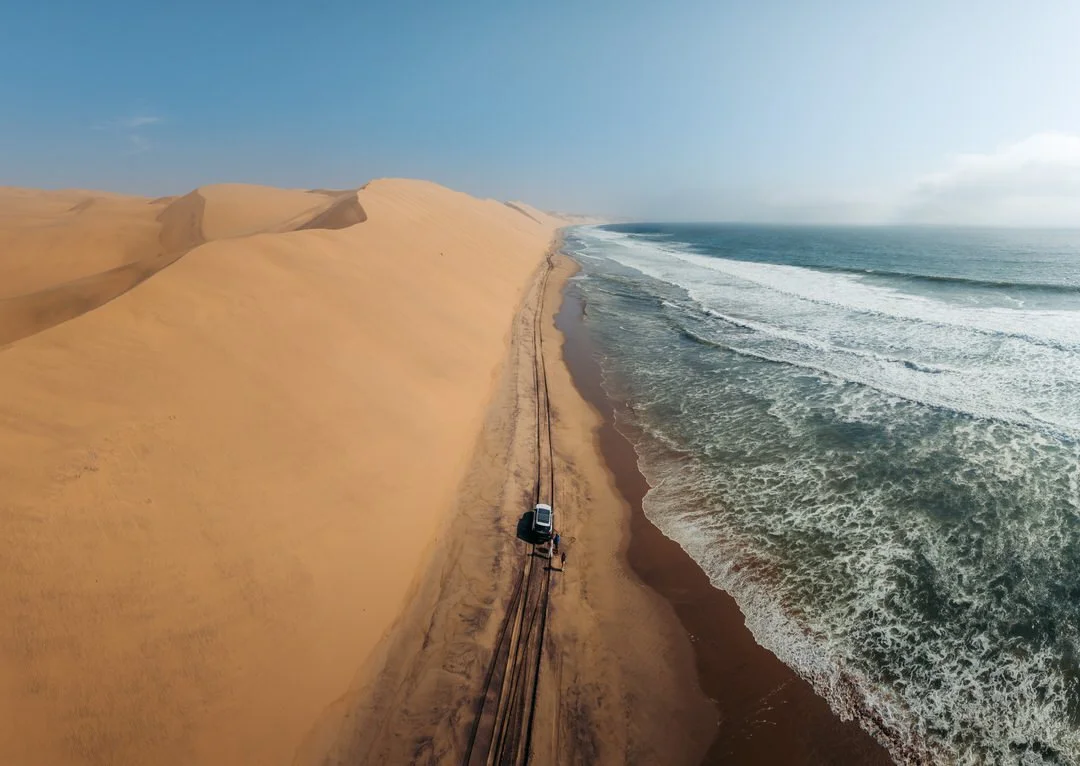 A vehicle traveling along tracks beside a sandy desert with high dunes on the left and an ocean with waves on the right.