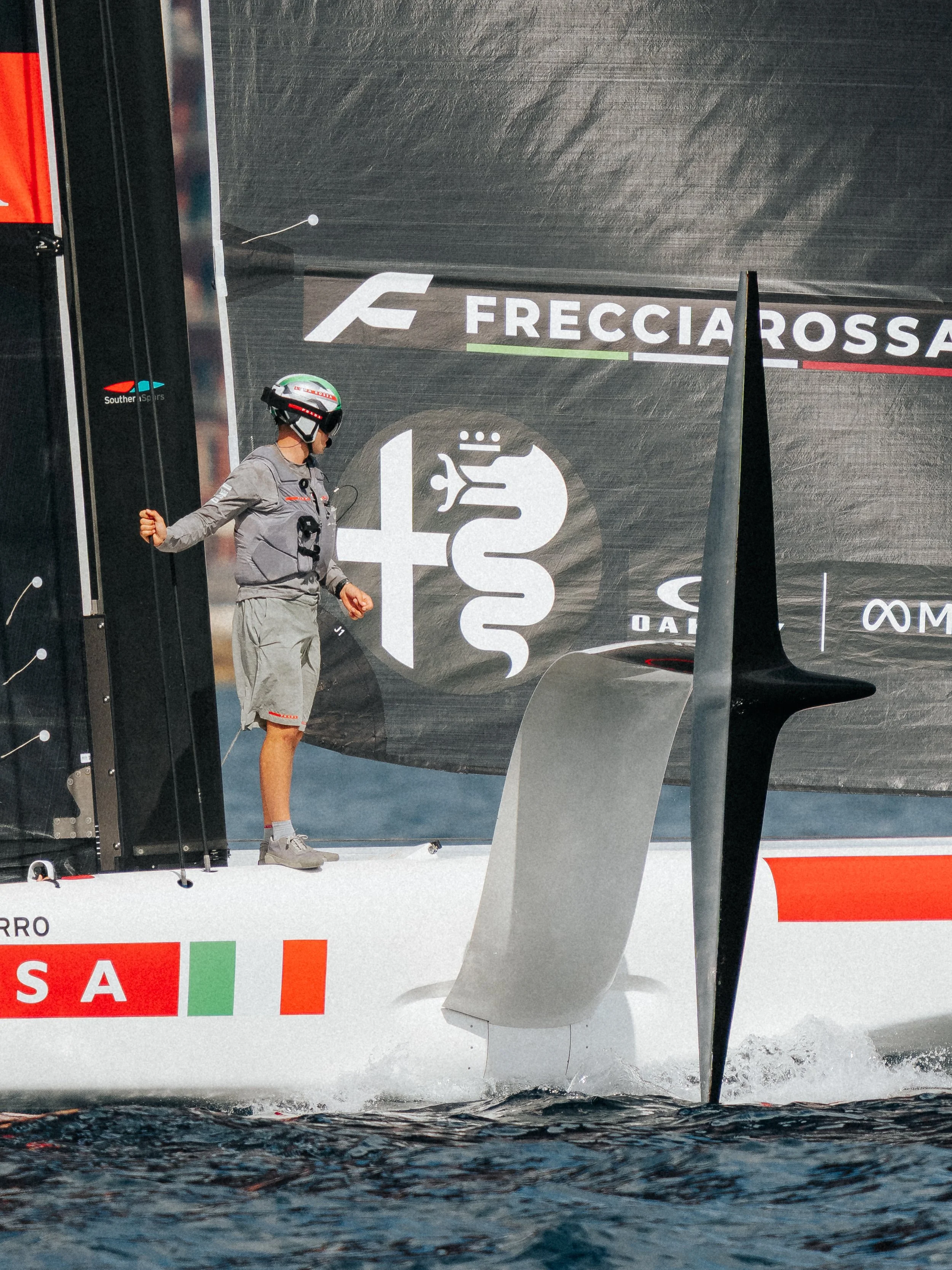 A person in racing gear and a helmet standing on a racing yacht deck near the sail with logos and sponsors in the background.