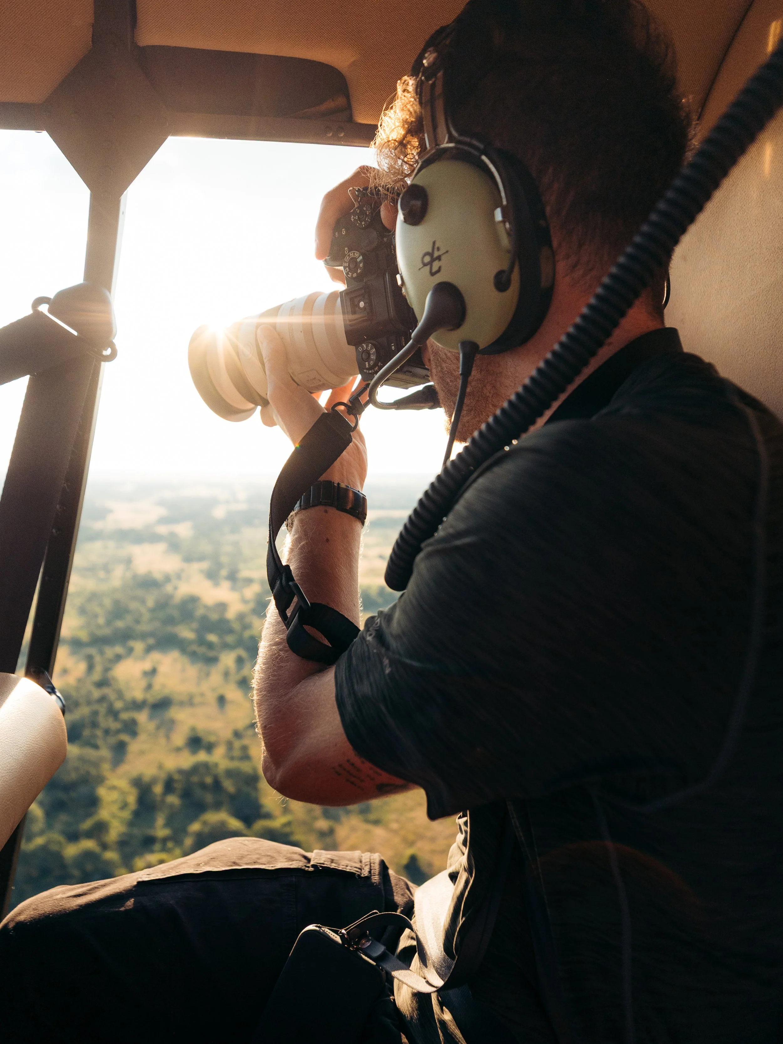 A man with curly hair wearing headphones and sunglasses, sitting in a helicopter, taking a photo with a camera, with the sun shining in the background over a landscape.