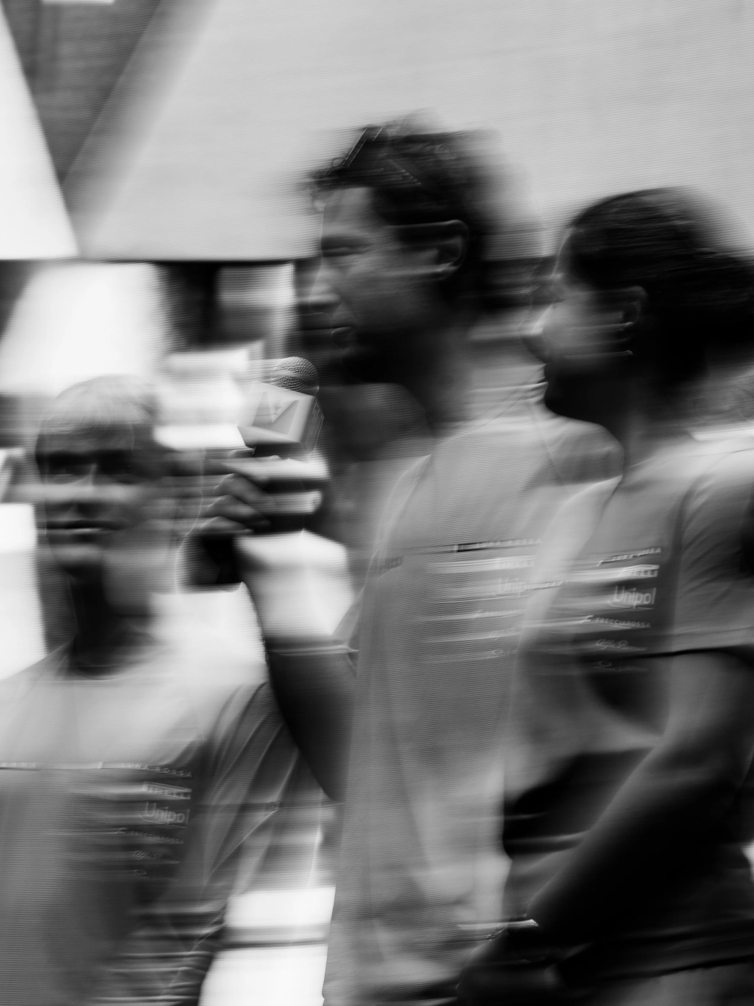 A blurred black and white photo of two people walking, one holding a microphone with a logo, and one person wearing glasses. The photo appears to be taken through a reflective or textured surface.