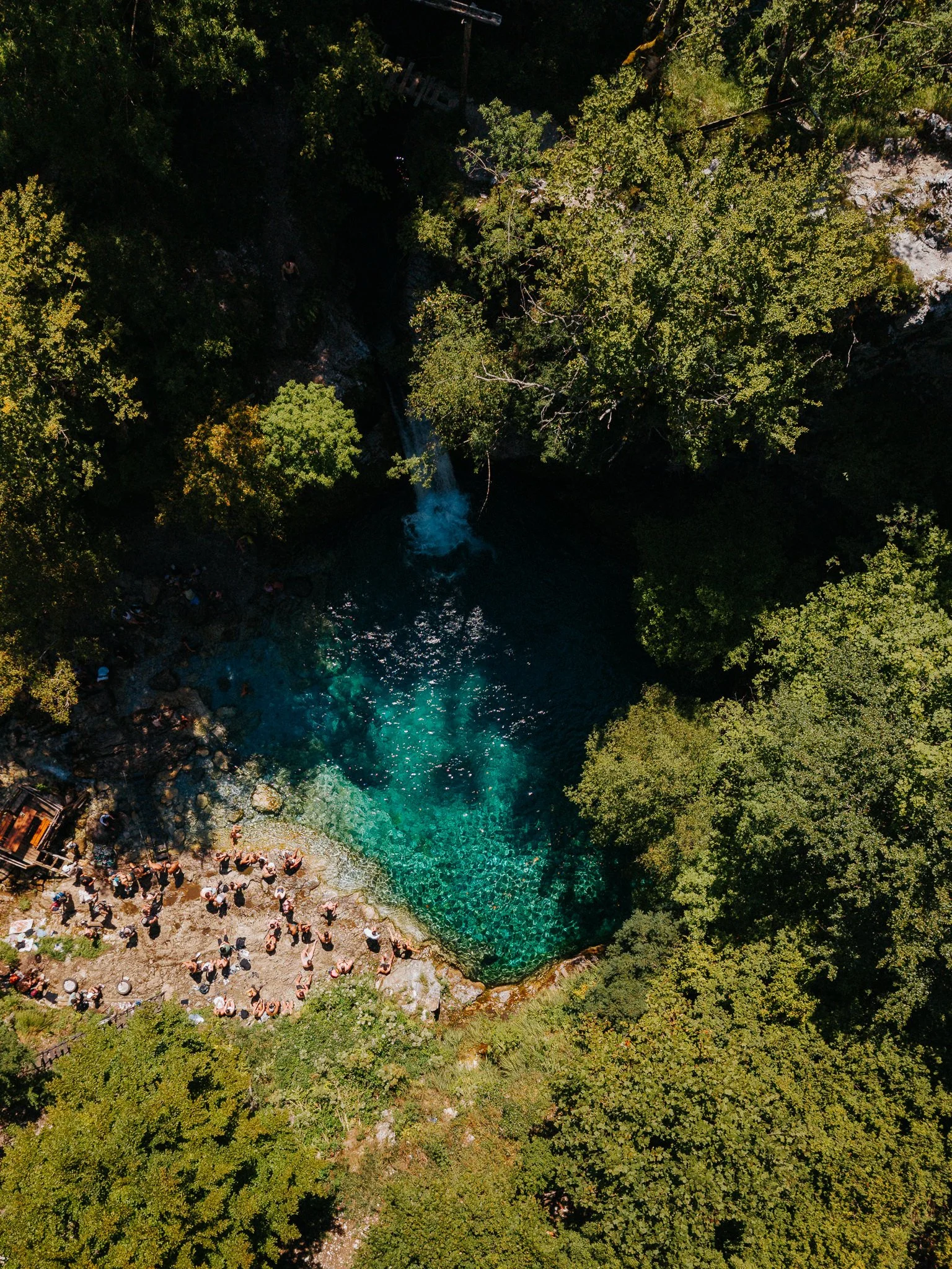 An aerial view of a natural swimming hole with a crowd of people gathered around the rocky shoreline, surrounded by lush green trees and a small waterfall cascading into the water.