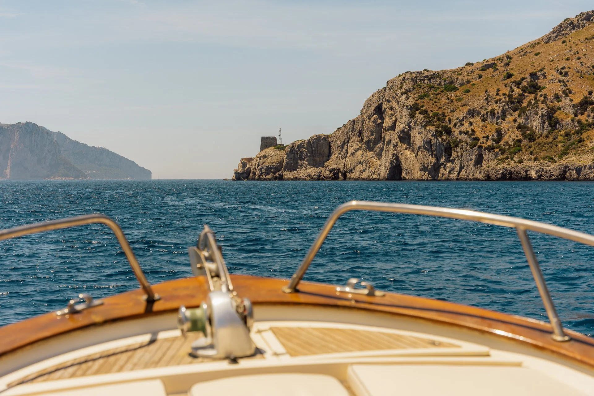 View of a coastline with rocky hills and a tower on the right, seen from the front of a boat on the water with part of the deck and railing visible in the foreground.