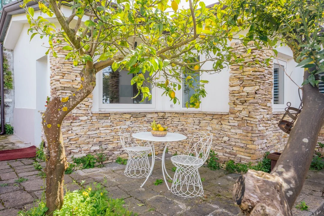 A cozy outdoor patio with a white metal table and two matching chairs, surrounded by greenery and trees, with a stone wall and house window in the background.