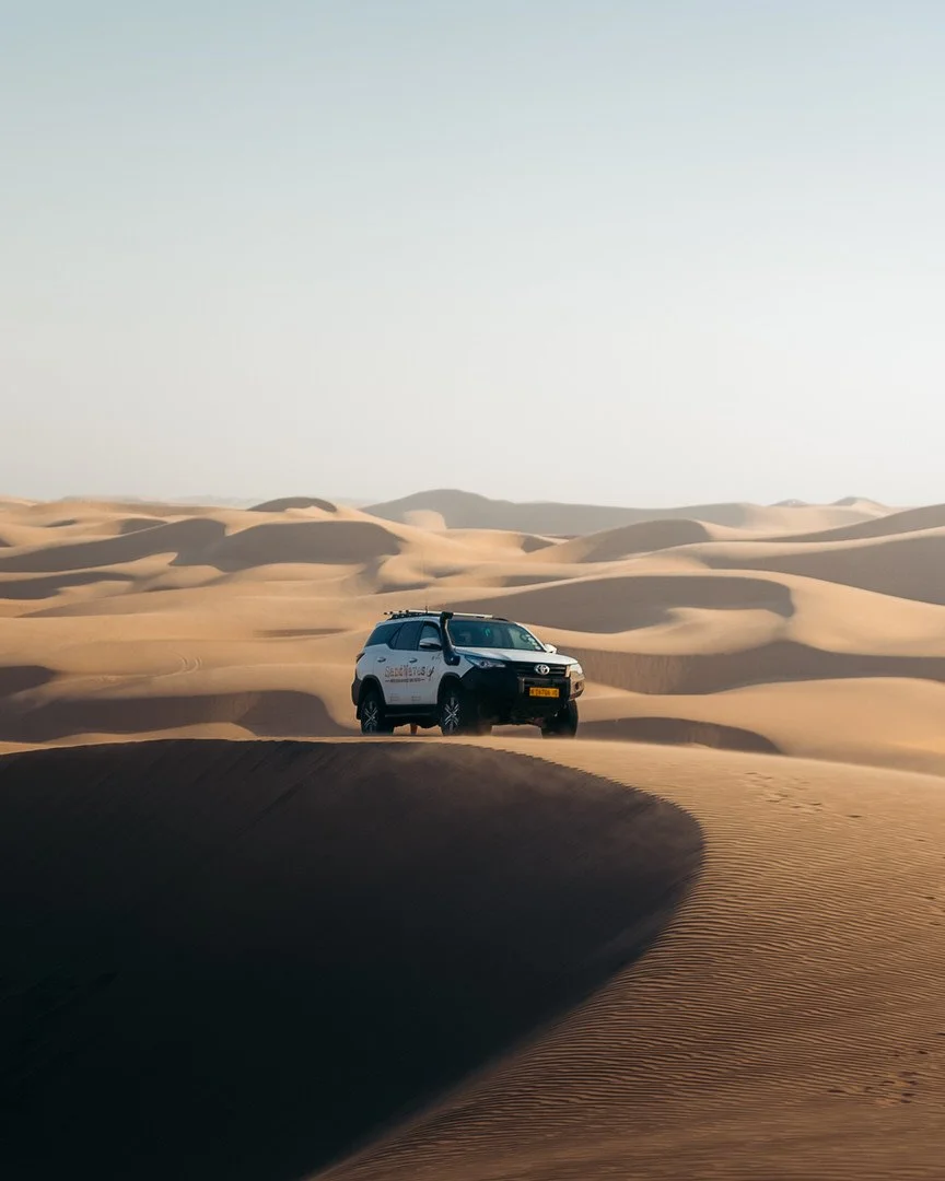 A white SUV driving on sand dunes in a desert landscape with clear sky.