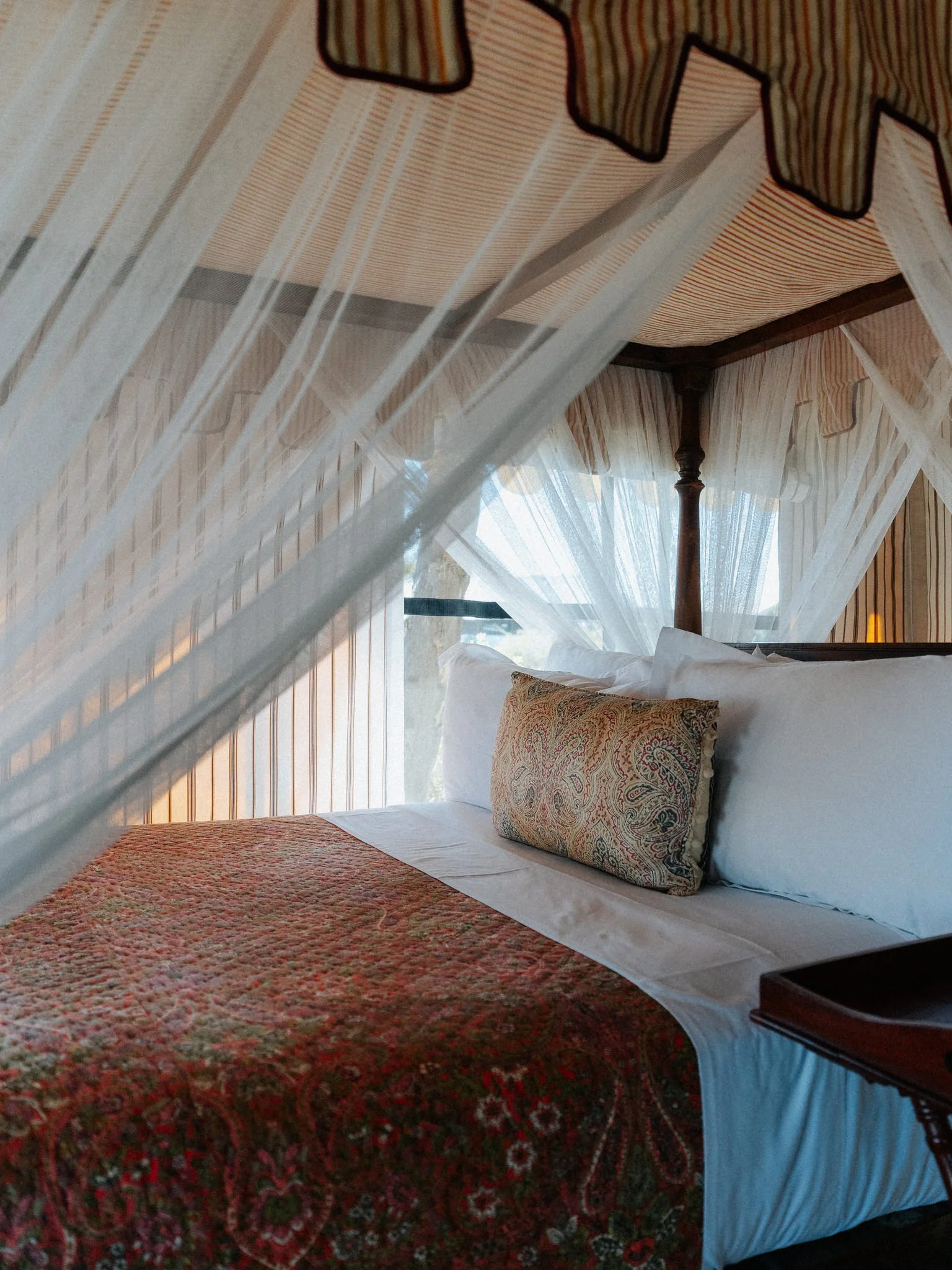View of a cozy hotel bedroom with a four-poster bed, white linens, a decorative pillow with a paisley pattern, and a red patterned bedspread. Sheer curtains hang around the bed, with sunlight filtering through a window.