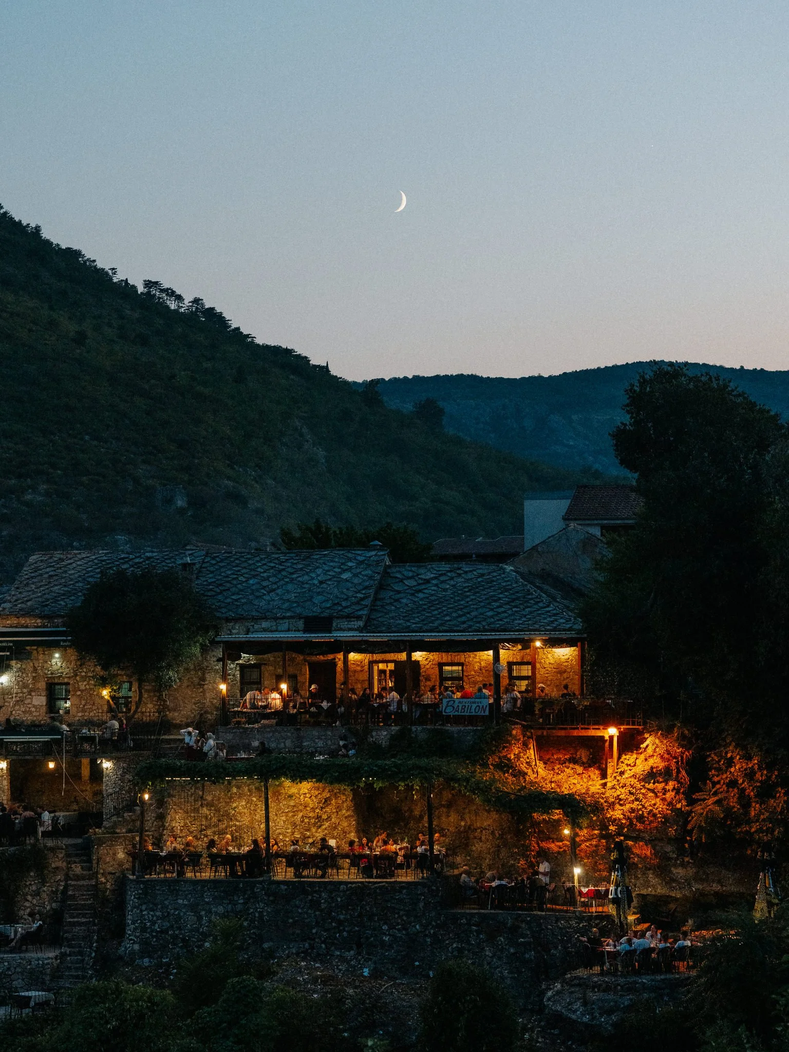 Night scene of a hillside restaurant with outdoor seating, illuminated by warm lights, under a sky with a crescent moon and mountains in the background.