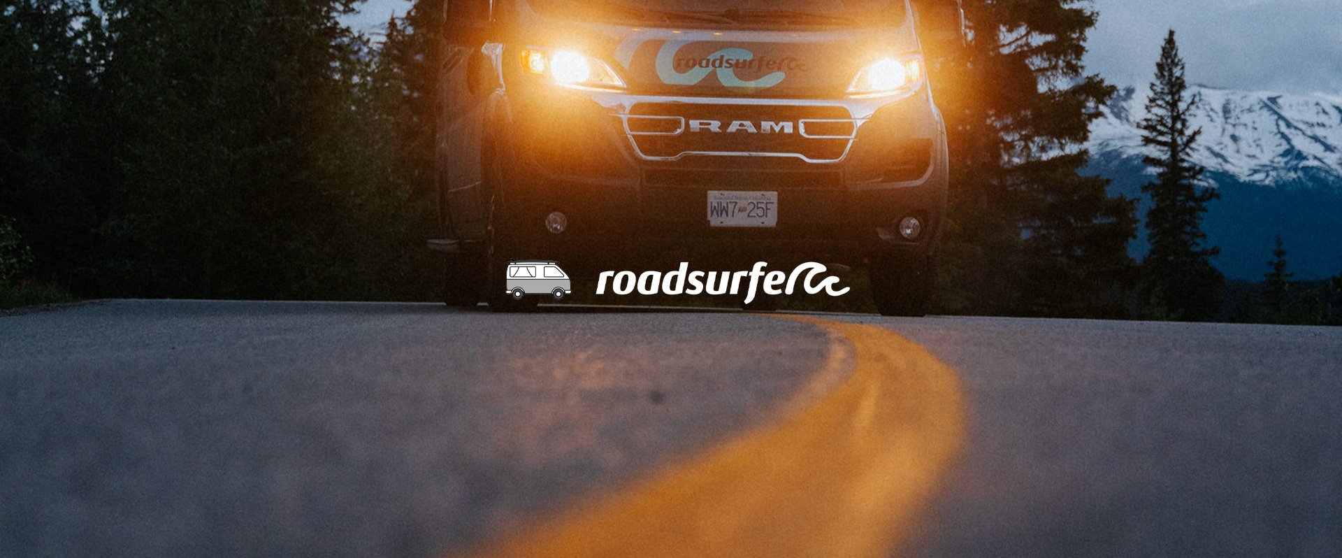 A truck with headlights on parked on a mountain road at dusk with snow-capped mountains and trees in the background. The image features the Roads of America logo with a small van icon.
