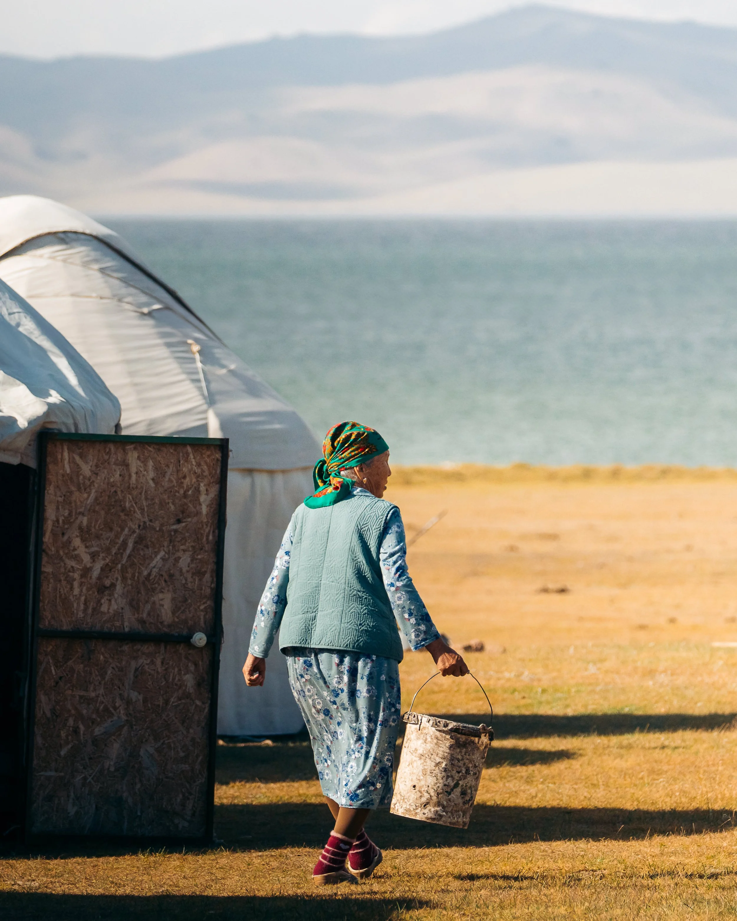 An elderly woman walking on a grassy beach holding a bucket, with tents and the ocean in the background.