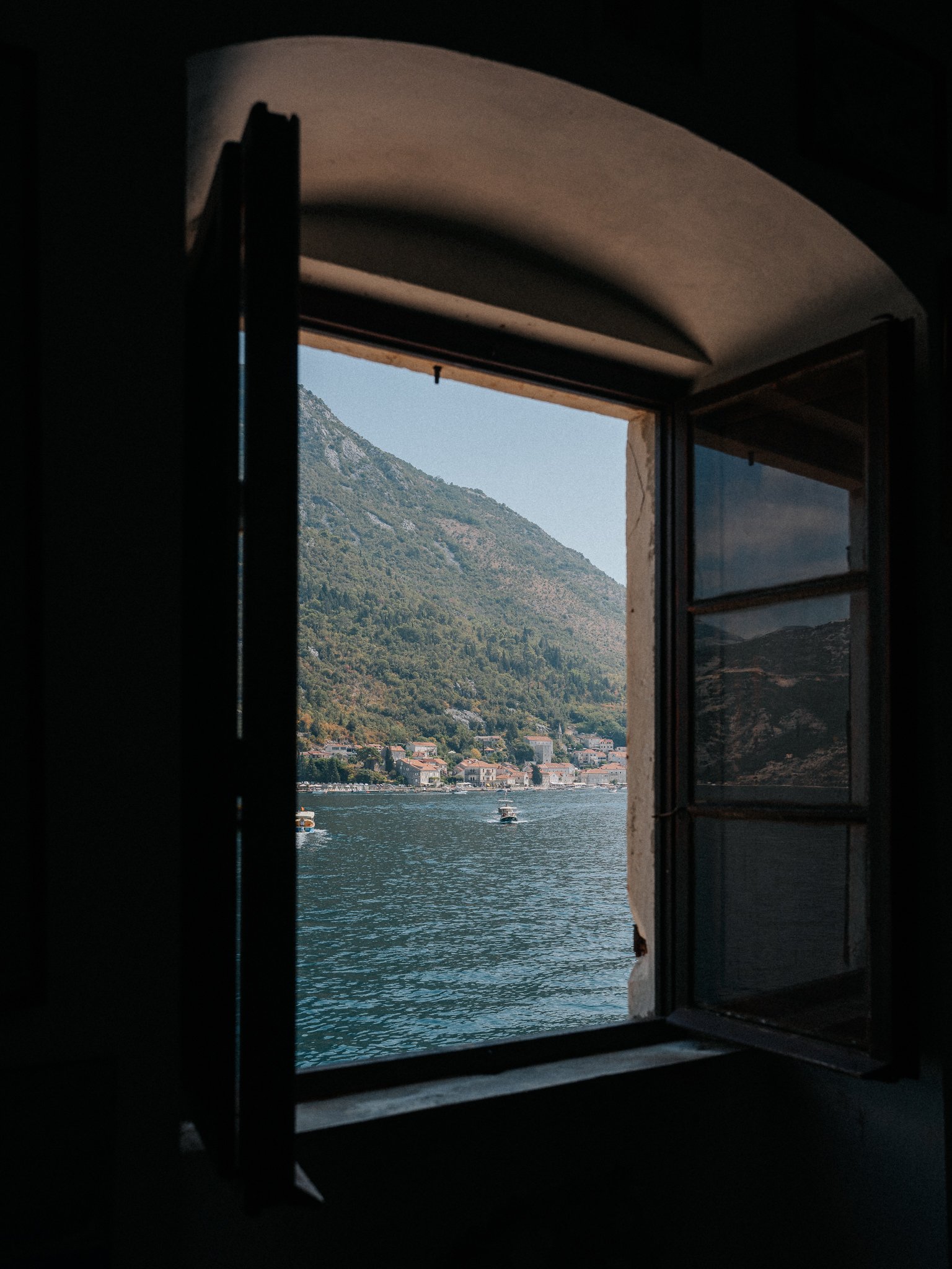 View through an open window of a river with boats, houses along the shore, and a mountain in the background under a clear sky.
