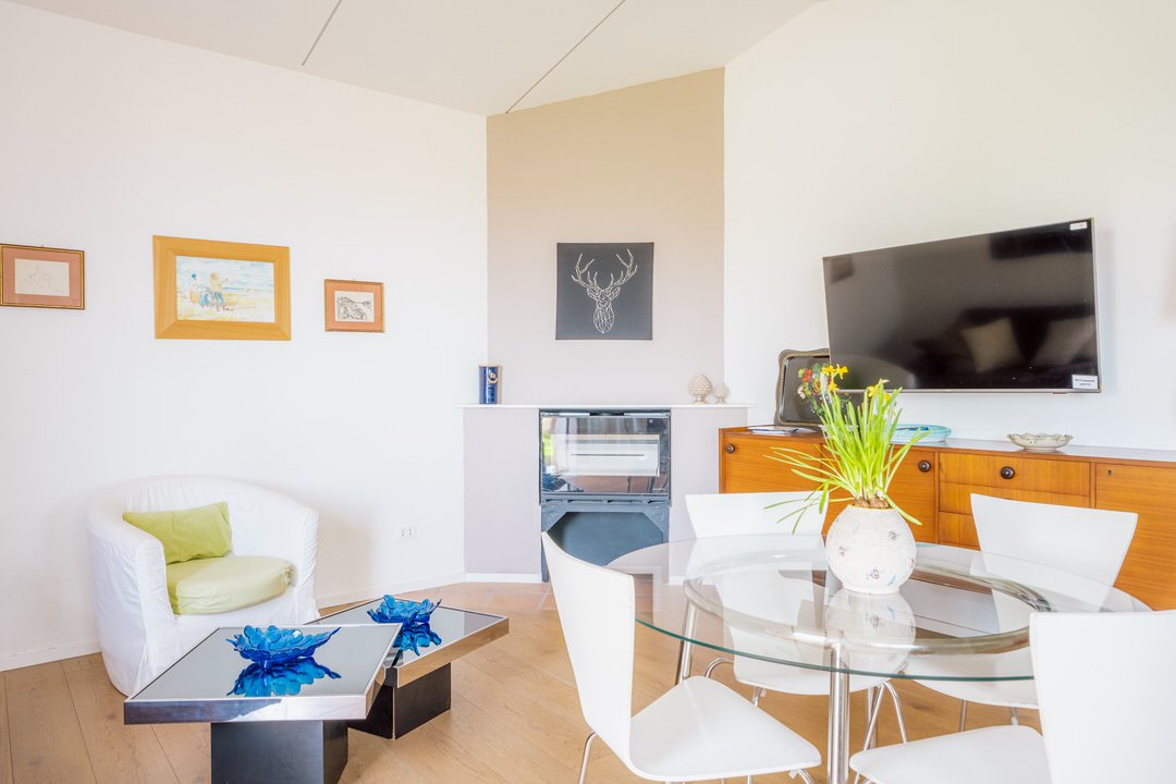 Living room with white walls, a white armchair with a green cushion, a modern glass-top coffee table with blue decorative accents, a round glass dining table with white chairs, a wooden sideboard, a flat-screen TV, and wall art including a deer sketc