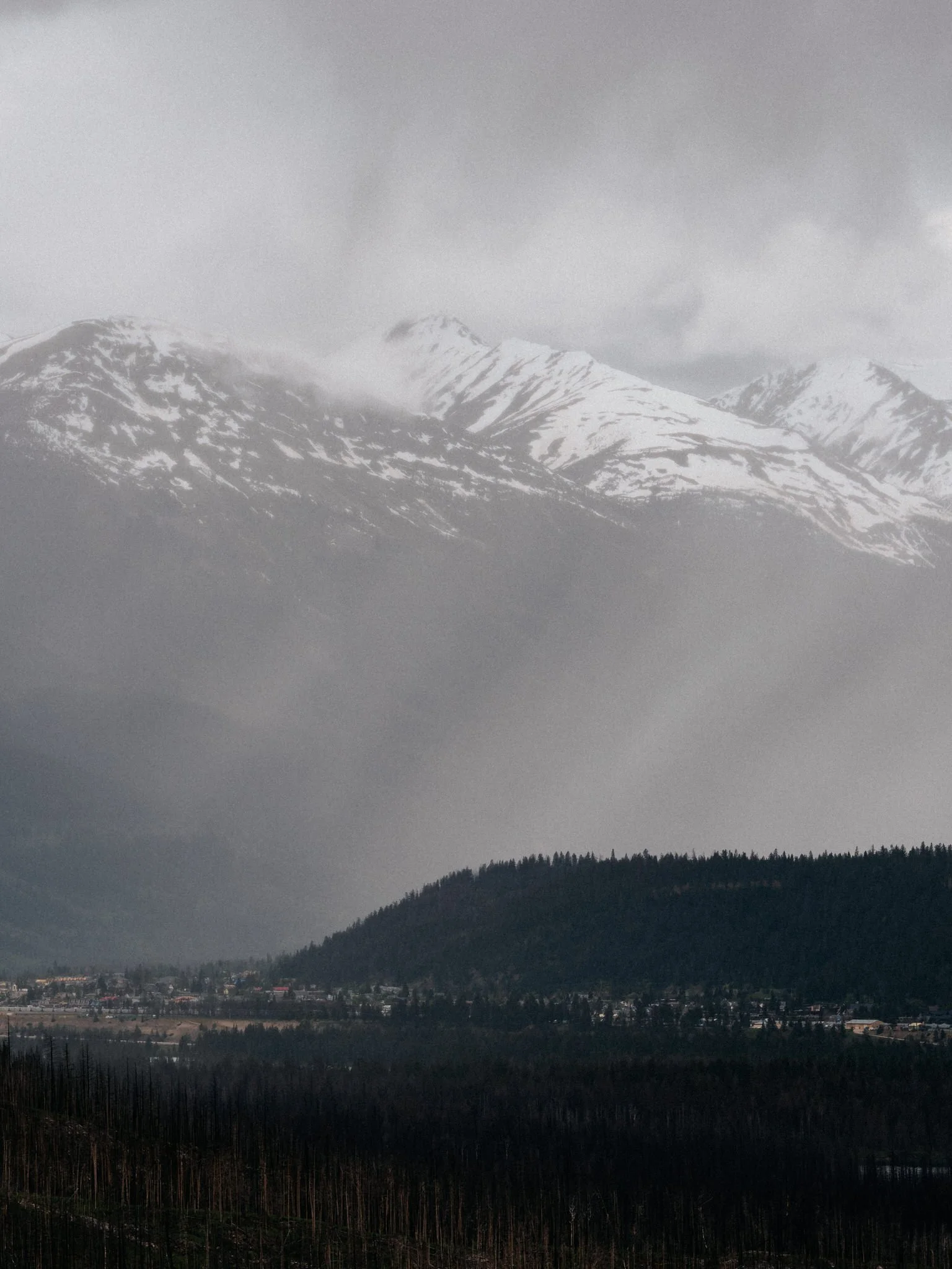 Snow-covered mountain peaks under a cloudy sky, with a forested hillside and small town at the base.