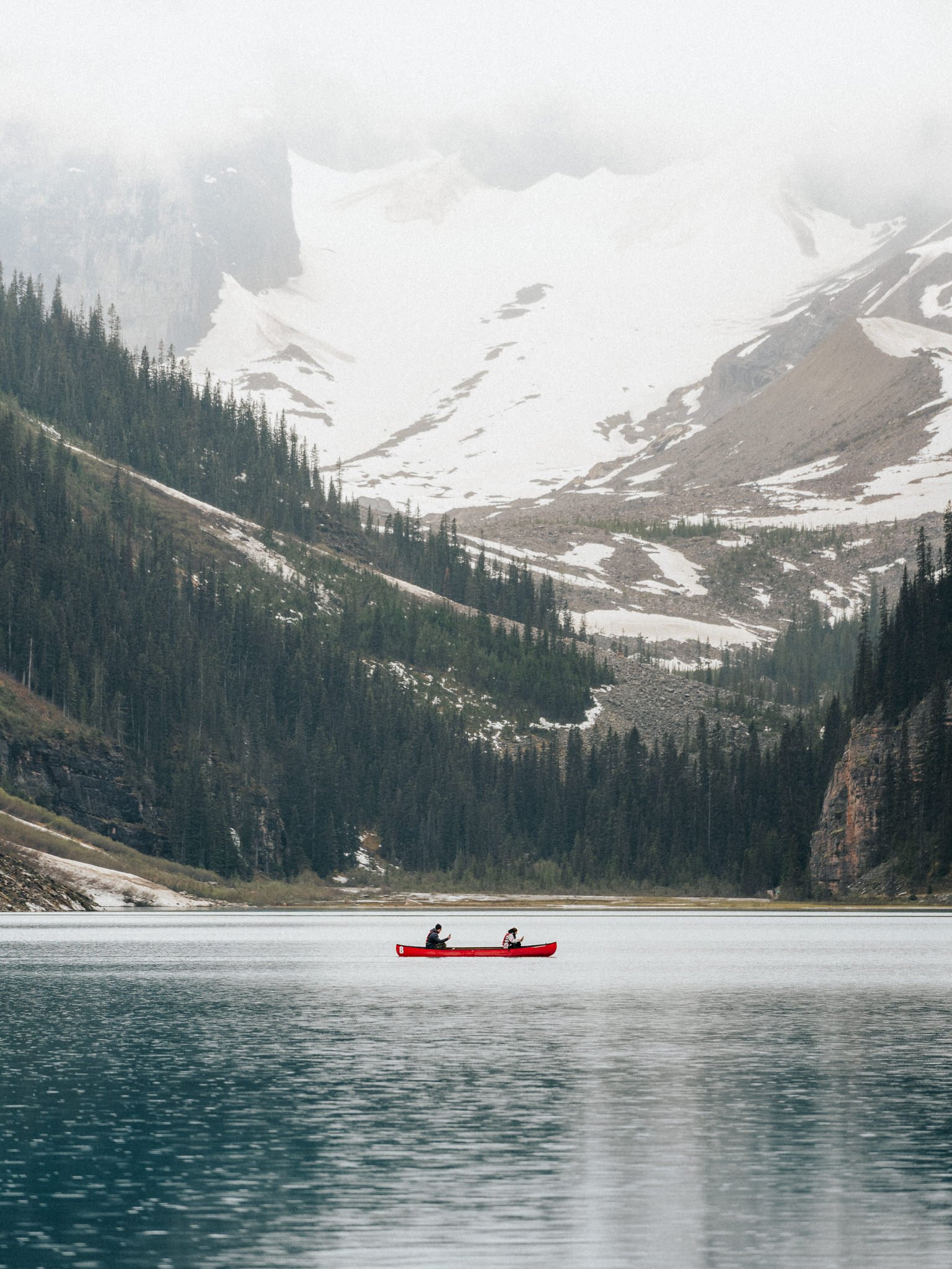 Two people paddling a red canoe on a calm lake surrounded by mountainous terrain with snow patches and forested slopes.