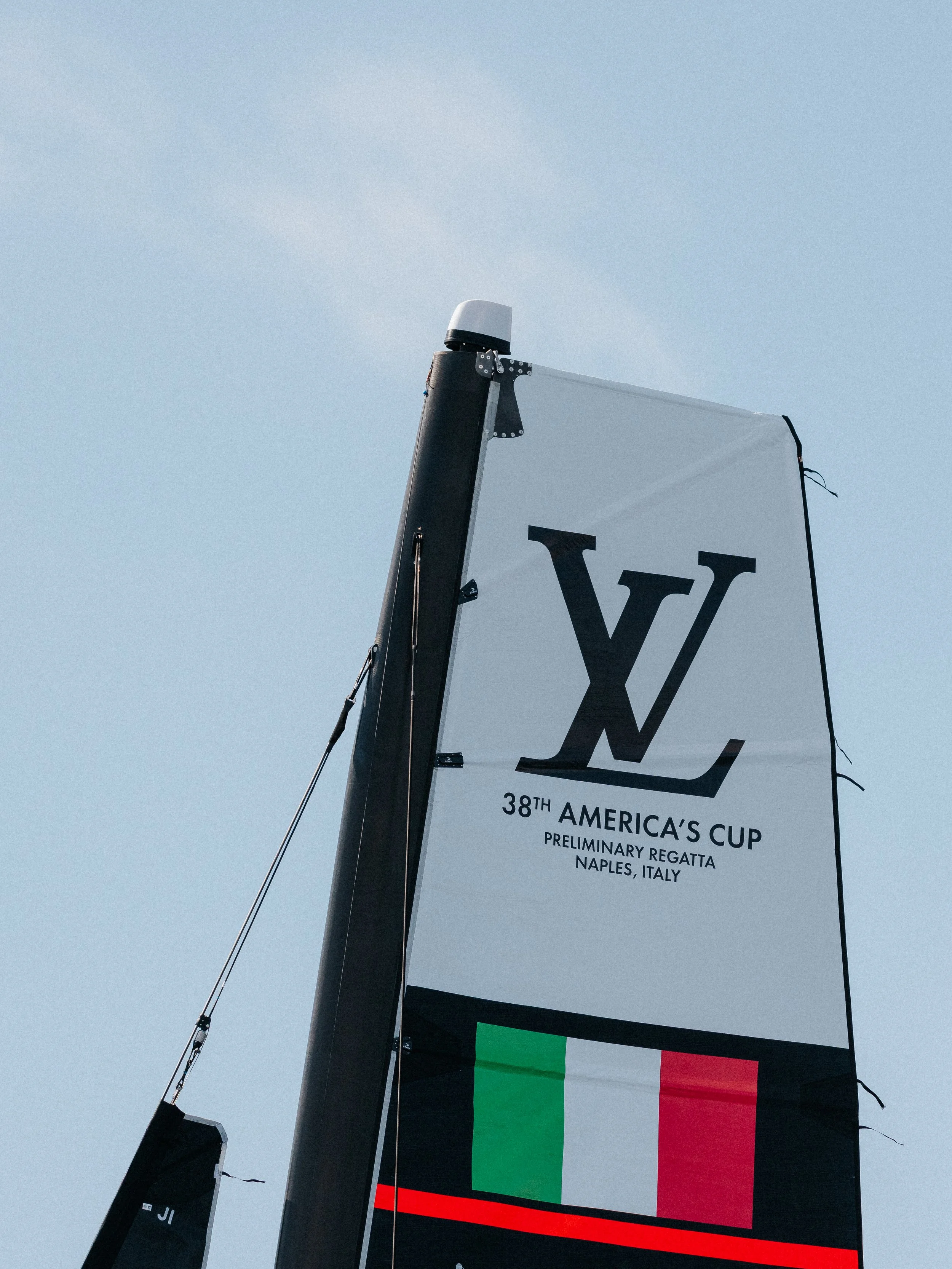 Sail banners on a boat mast at the 38th America's Cup preliminary regatta in Naples, Italy, featuring the Louis Vuitton logo and the Italian flag.