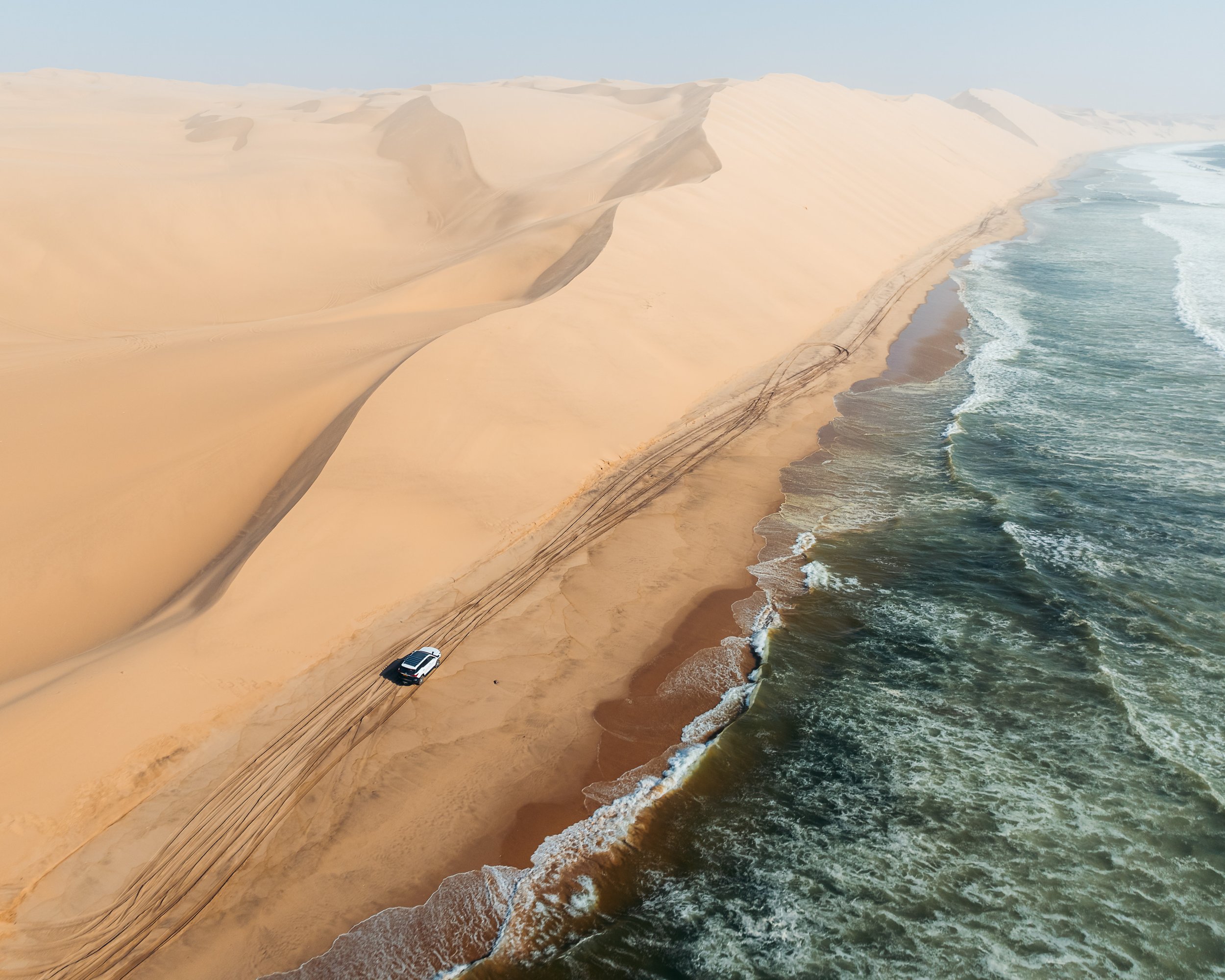 A car driving on a sandy beach next to crashing ocean waves with high sand dunes in the background.