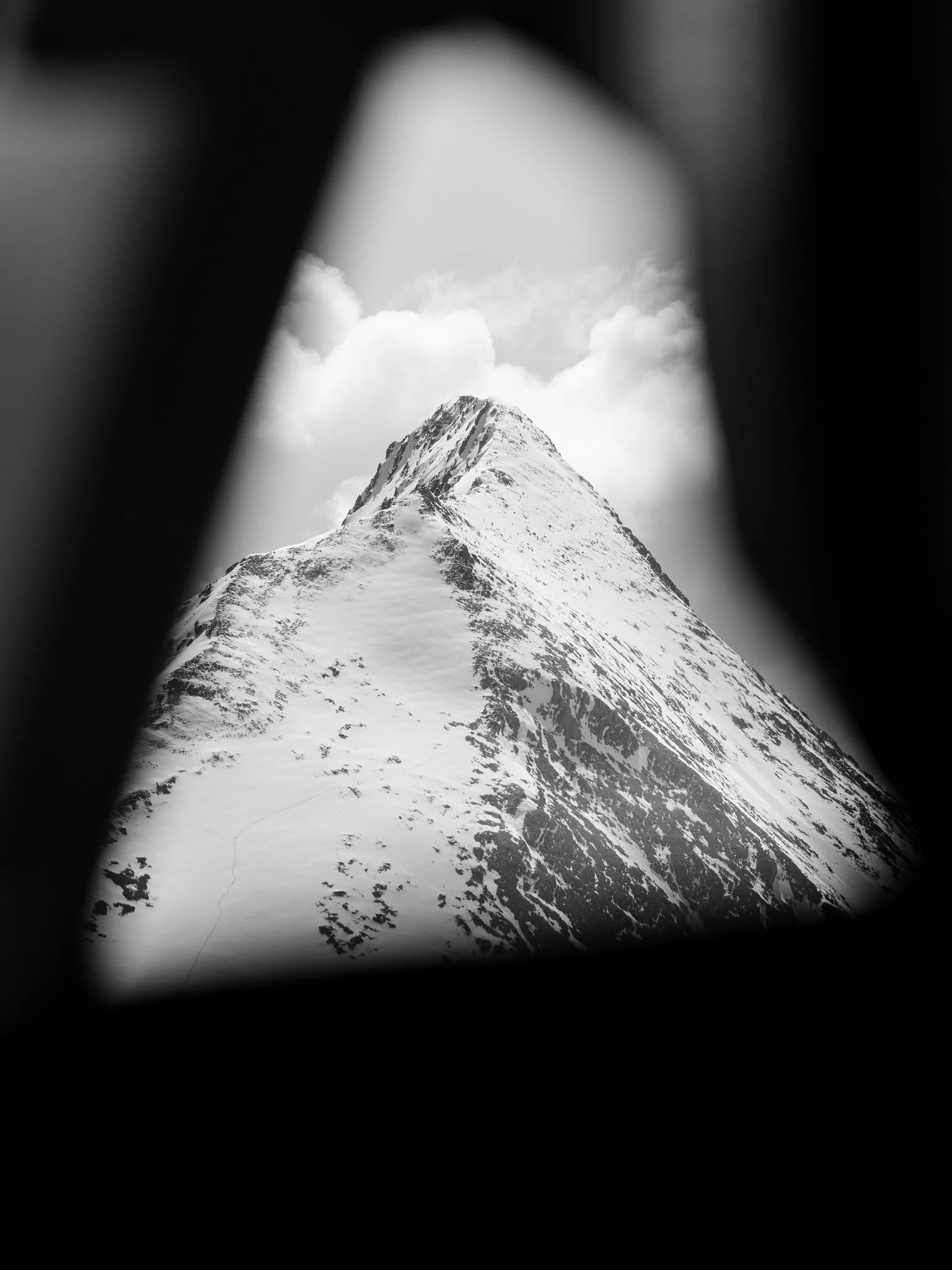 Black and white photo of a snow-covered mountain peak seen through a narrow, dark opening.