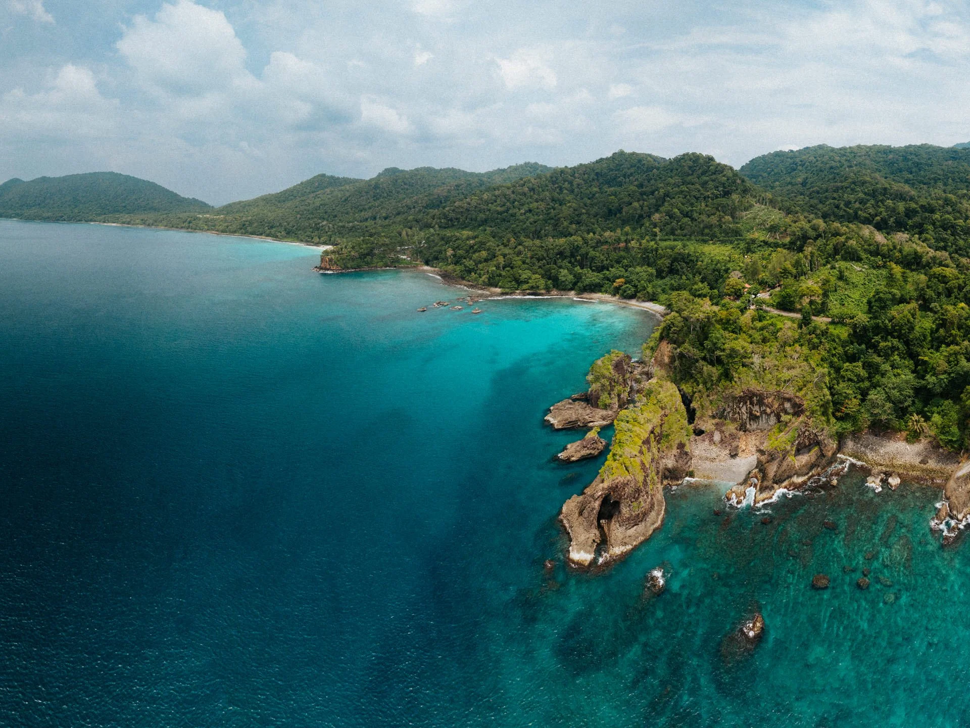 Aerial view of a tropical coastline with green hills, a clear blue ocean, and rocky cliffs