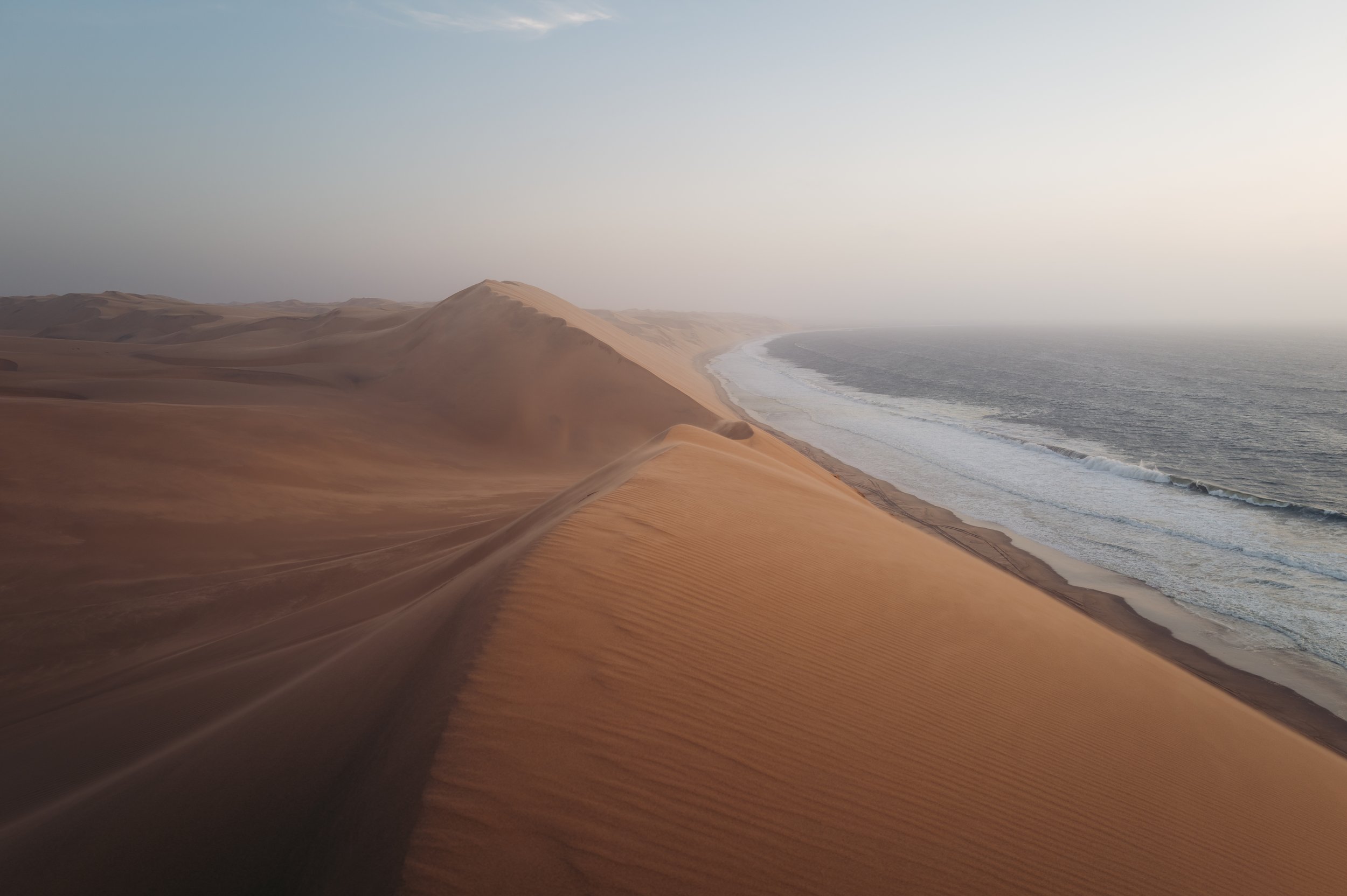 Aerial view of large sand dunes along a coastline with ocean waves, during sunset or sunrise.