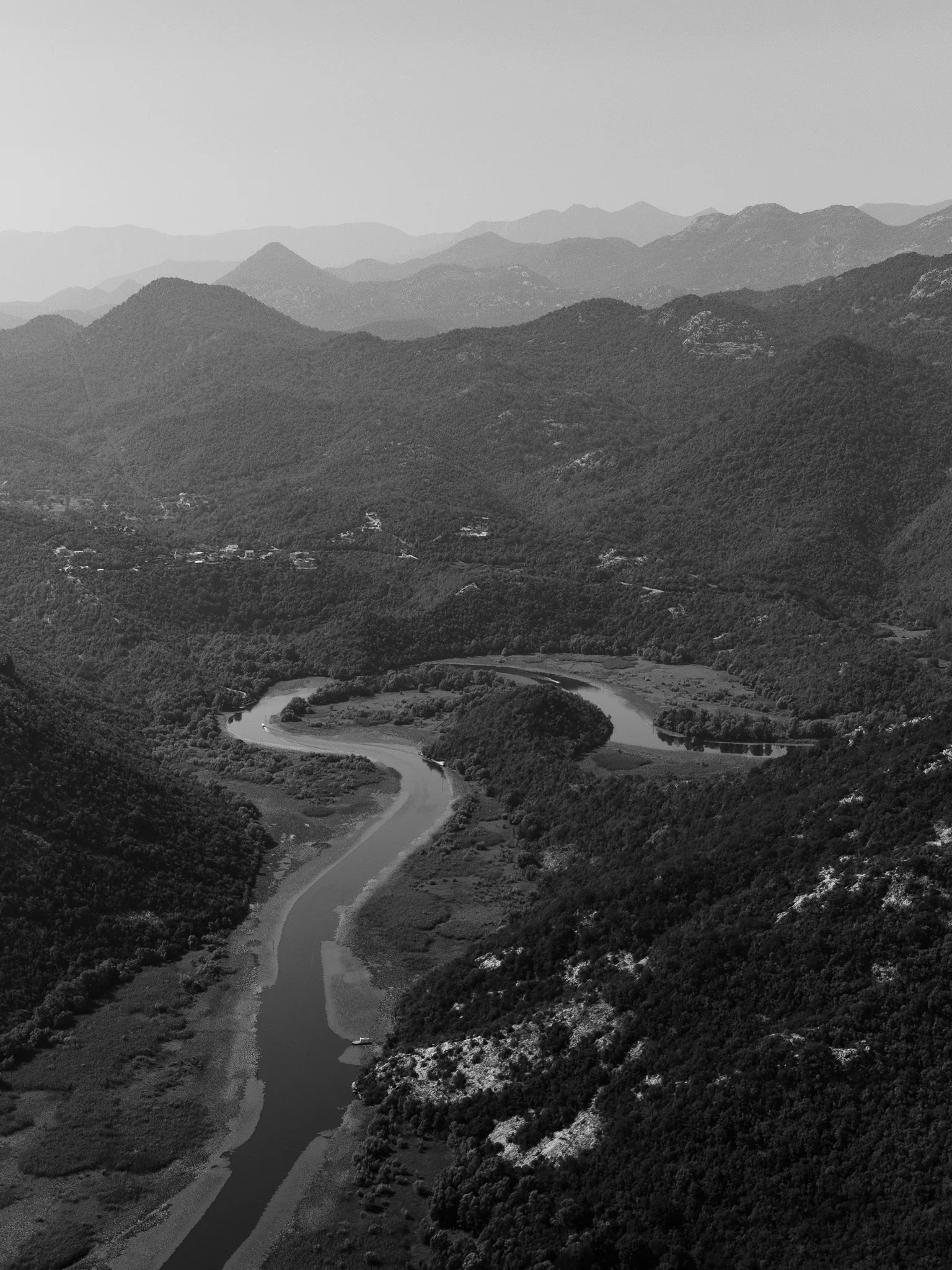 A black and white aerial view of a winding river flowing through a lush, forested valley surrounded by mountainous terrain.