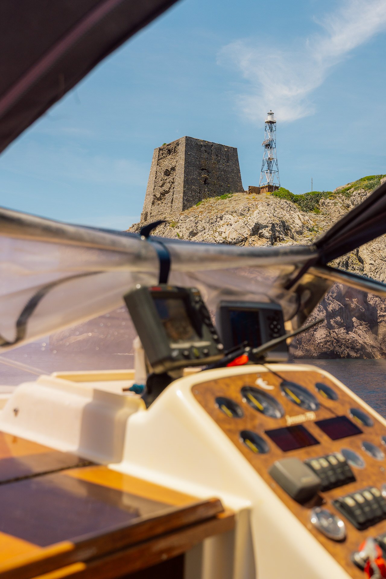 View from inside a boat with navigation equipment, looking out at a rocky hill with an old stone fort and a lighthouse tower against a blue sky.