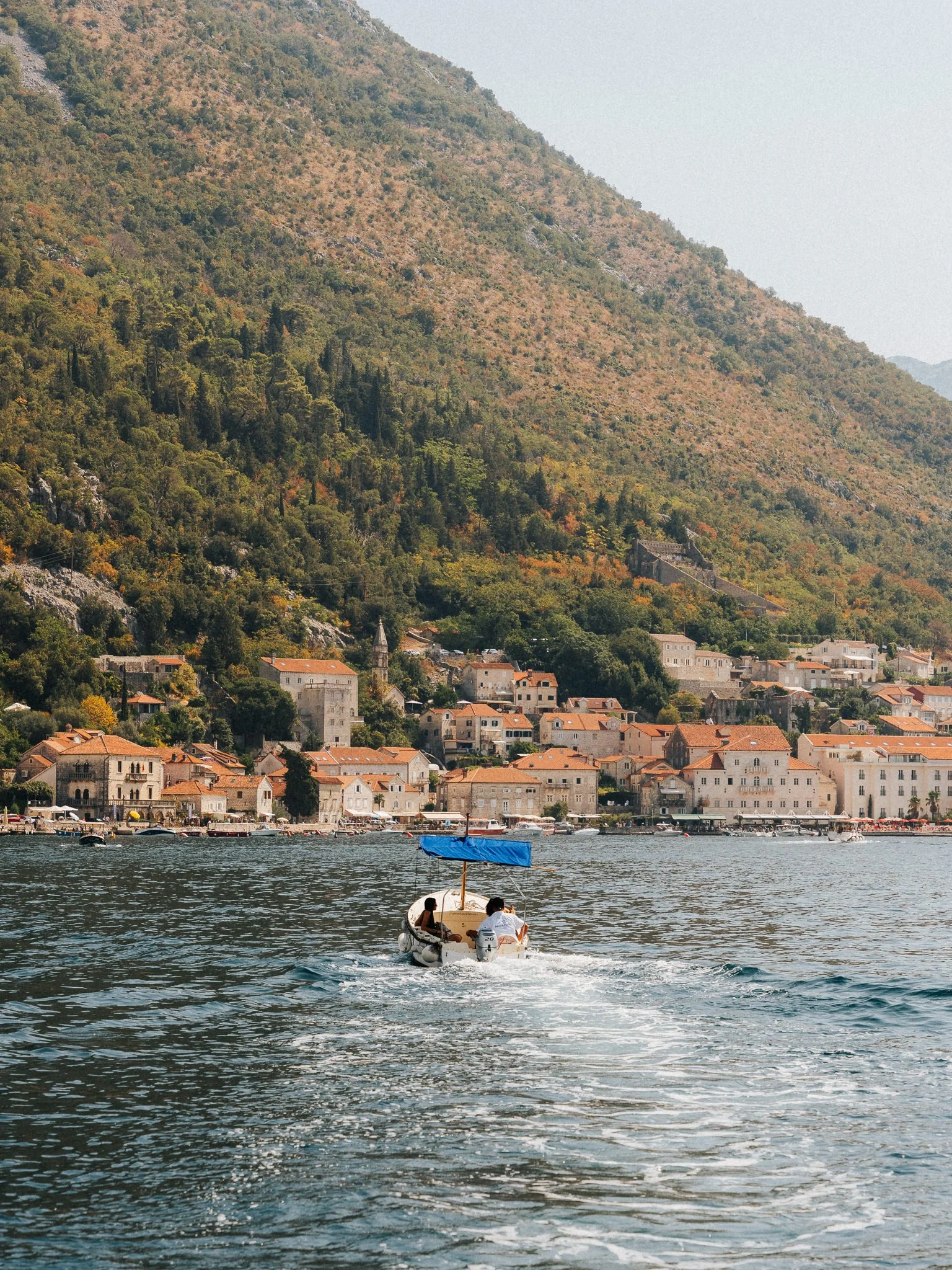 A small boat with two people on the water, near a coastal town with red roofs and historic buildings, backed by a mountain covered with trees.